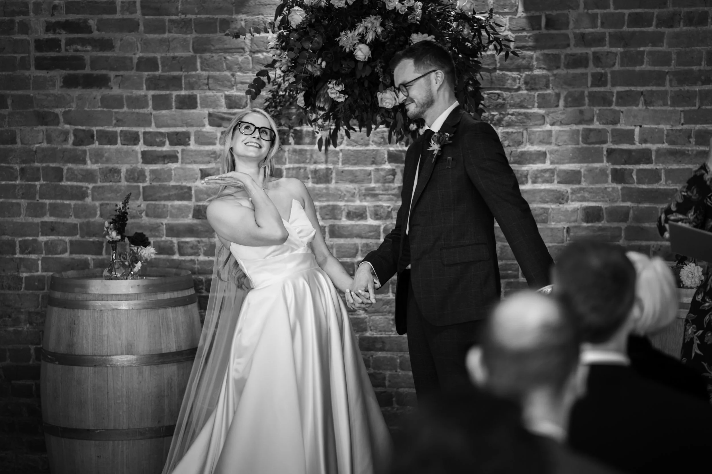 A black-and-white photo of a wedding ceremony where a smiling bride and groom hold hands in front of a brick wall with floral decorations. The bride is wearing a strapless wedding dress and glasses, and the groom is in a dark suit with glasses. Guest