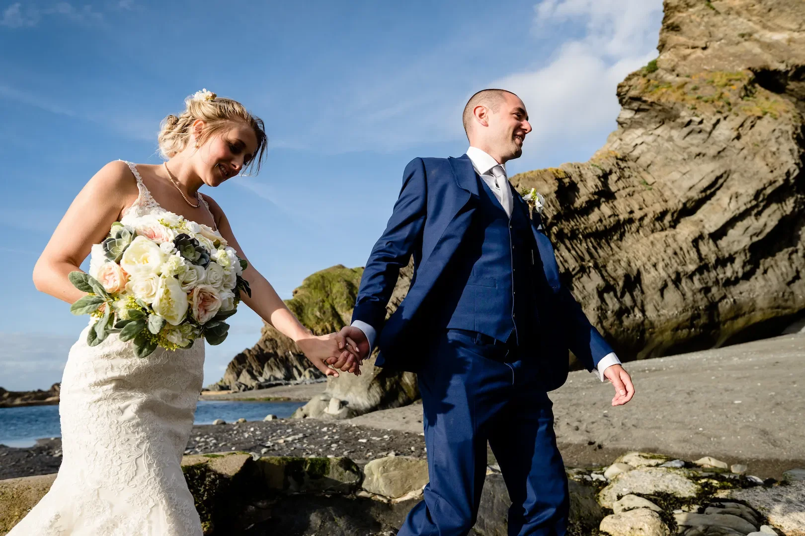 A bride and groom holding hands on a rocky beach with large cliffs in the background during a sunny day.