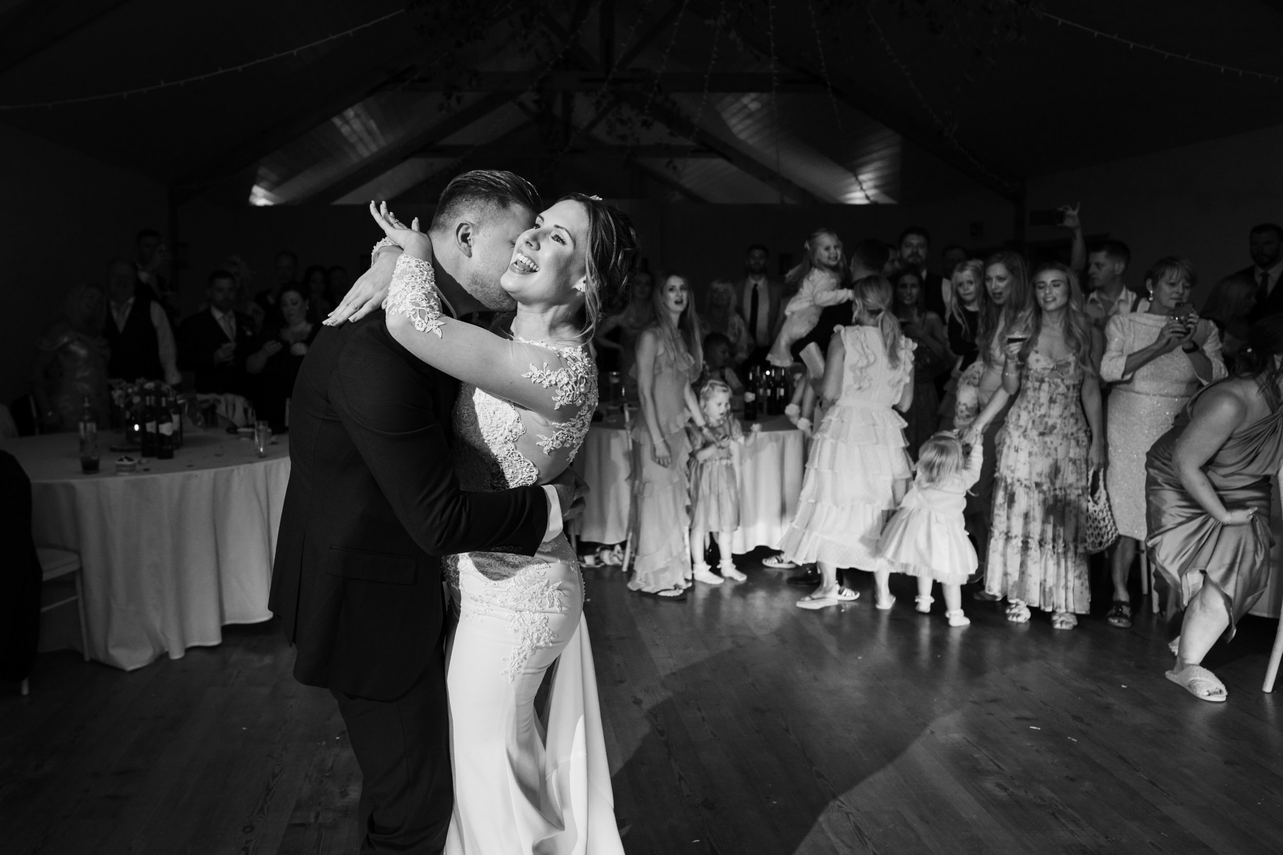 A bride and groom dancing closely at their wedding reception, surrounded by guests including children, in a festive indoor setting.