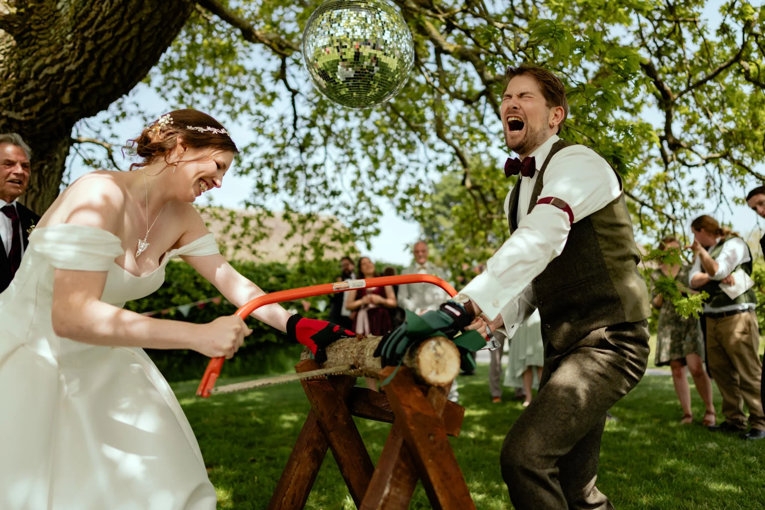A bride and groom enthusiastically sawing a log together outside during a wedding celebration, with onlookers in the background.