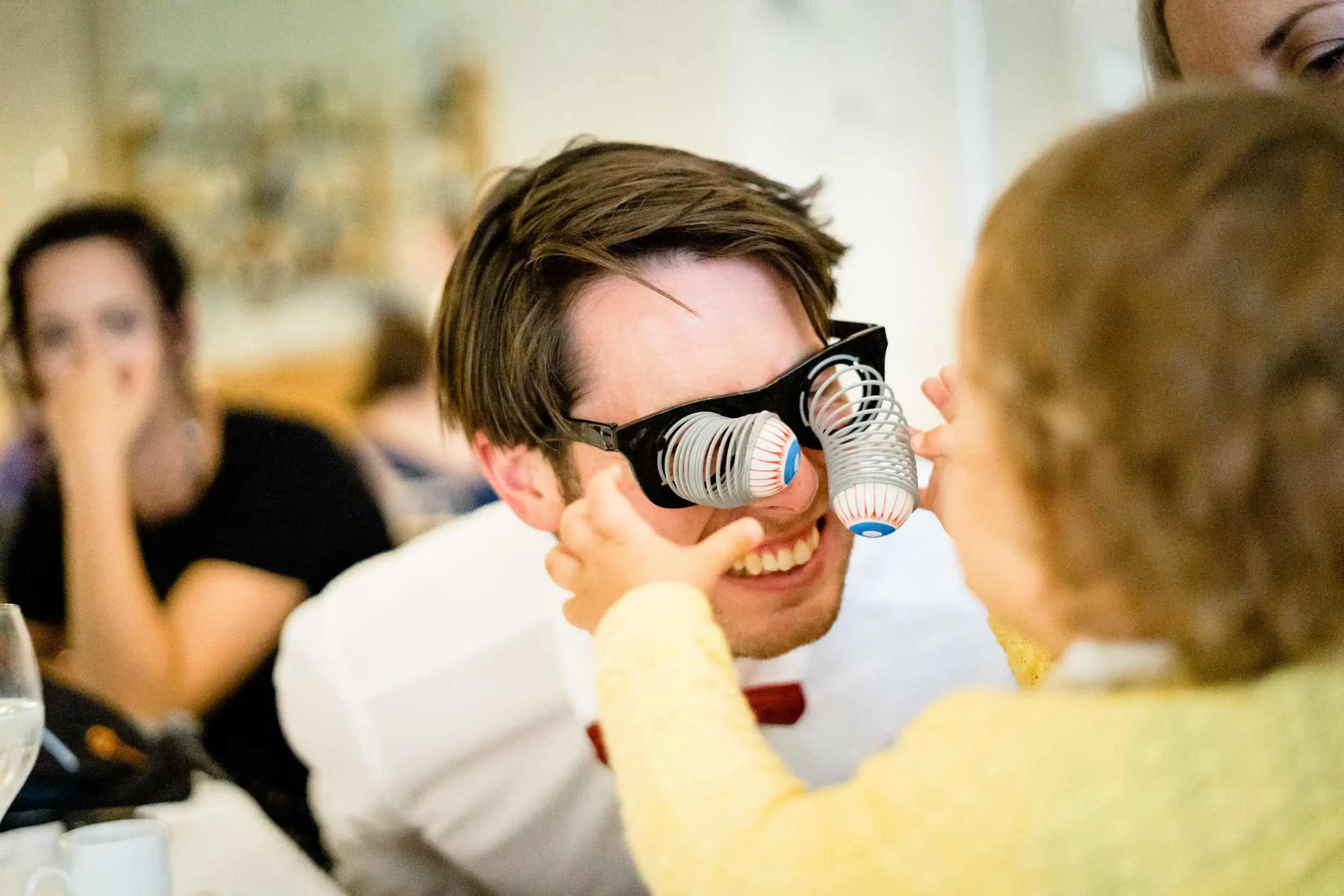 Person wearing novelty glasses with plastic eyeballs attached, smiling and playing with a child's face in a social gathering.