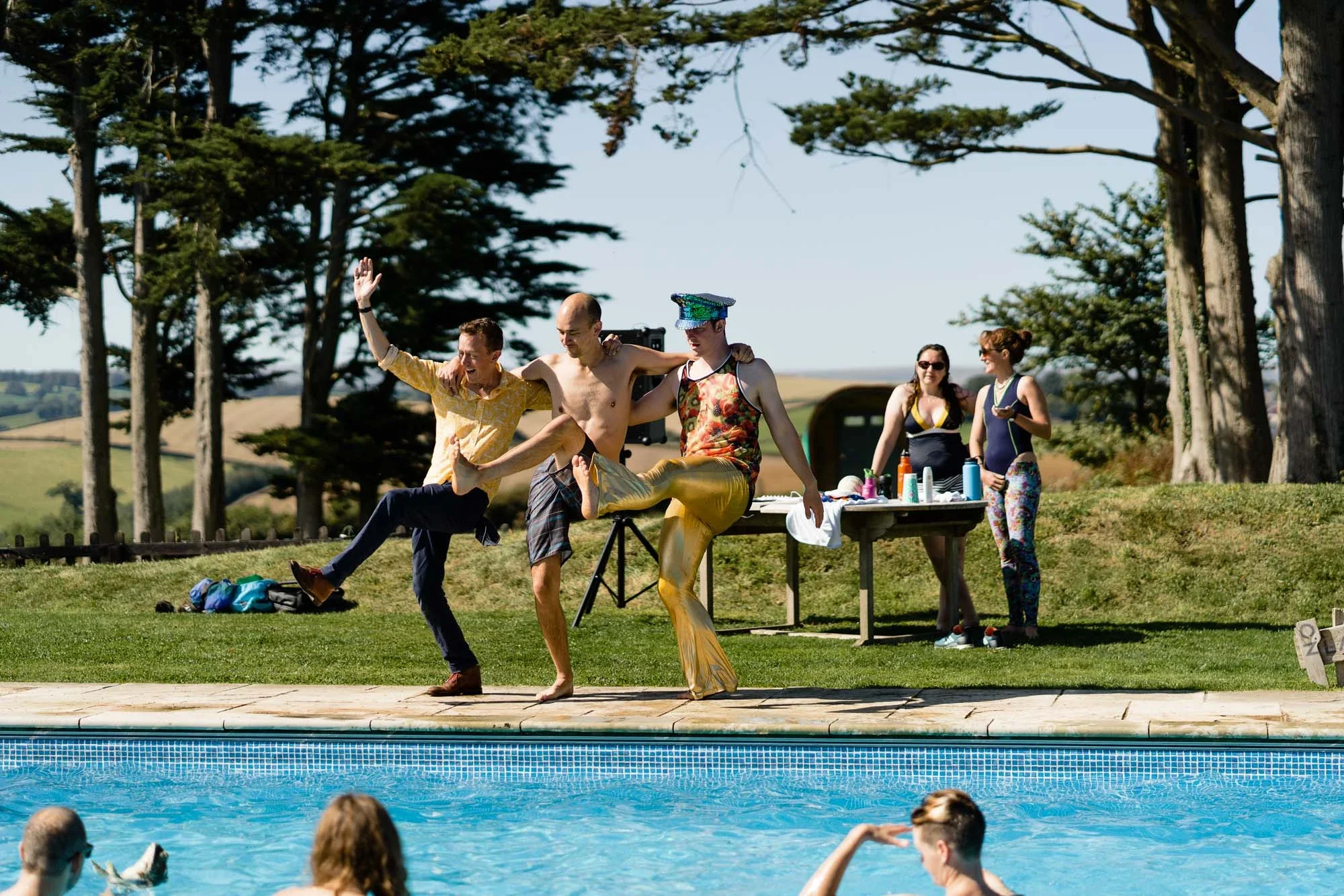 A group of people dressed in colorful outfits and engaging in a playful or dance activity by a swimming pool, with others watching and socializing nearby.