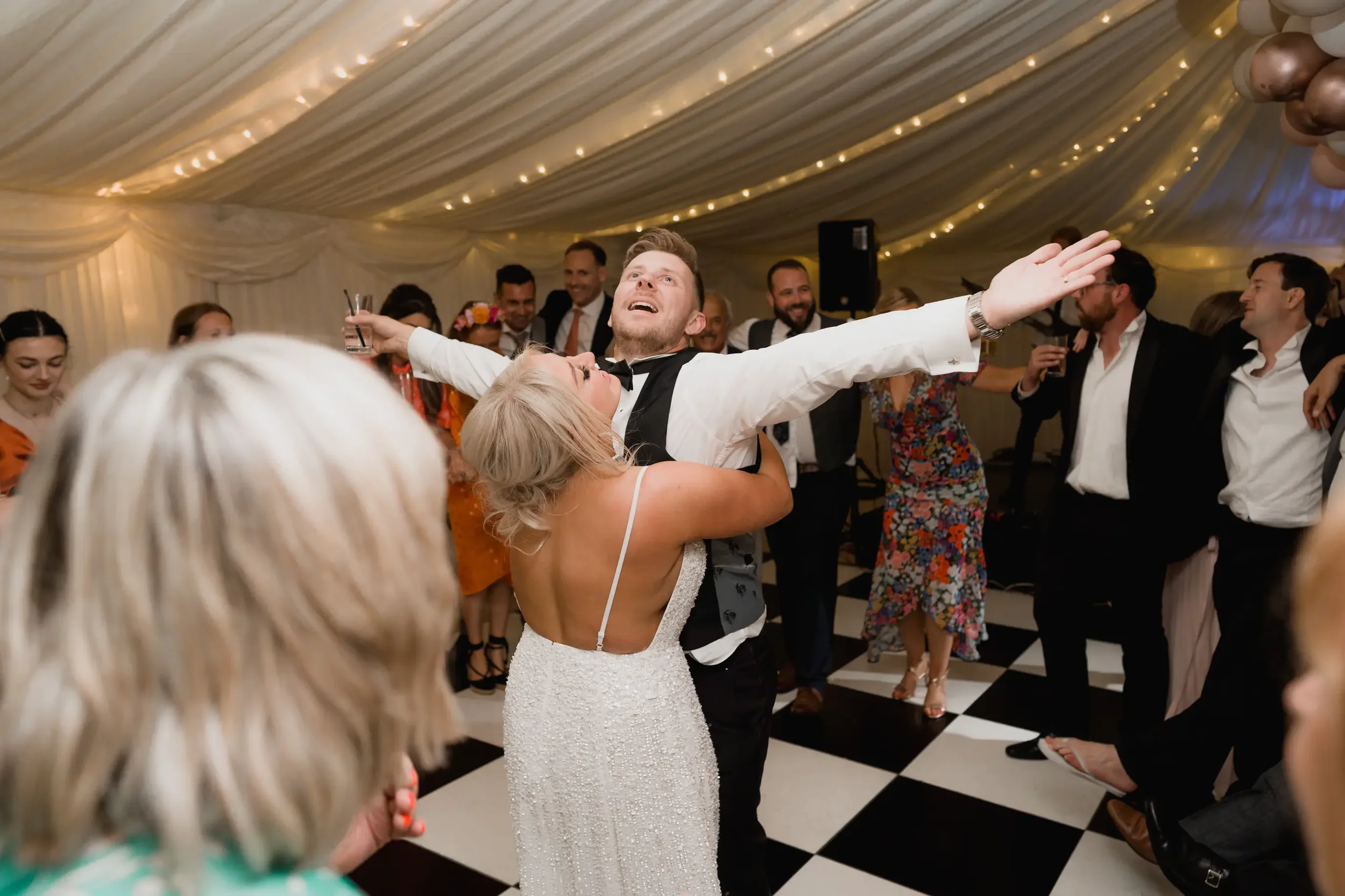 A groom dancing and joyfully lifting his bride during a celebration in a large decorated tent, surrounded by guests watching and smiling.