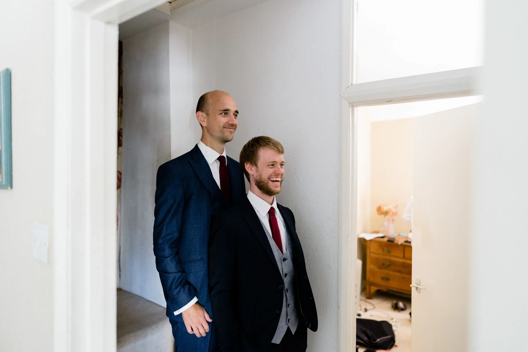 Two men dressed in suits standing in a doorway, smiling and looking at something off-camera, with a room visible in the background.