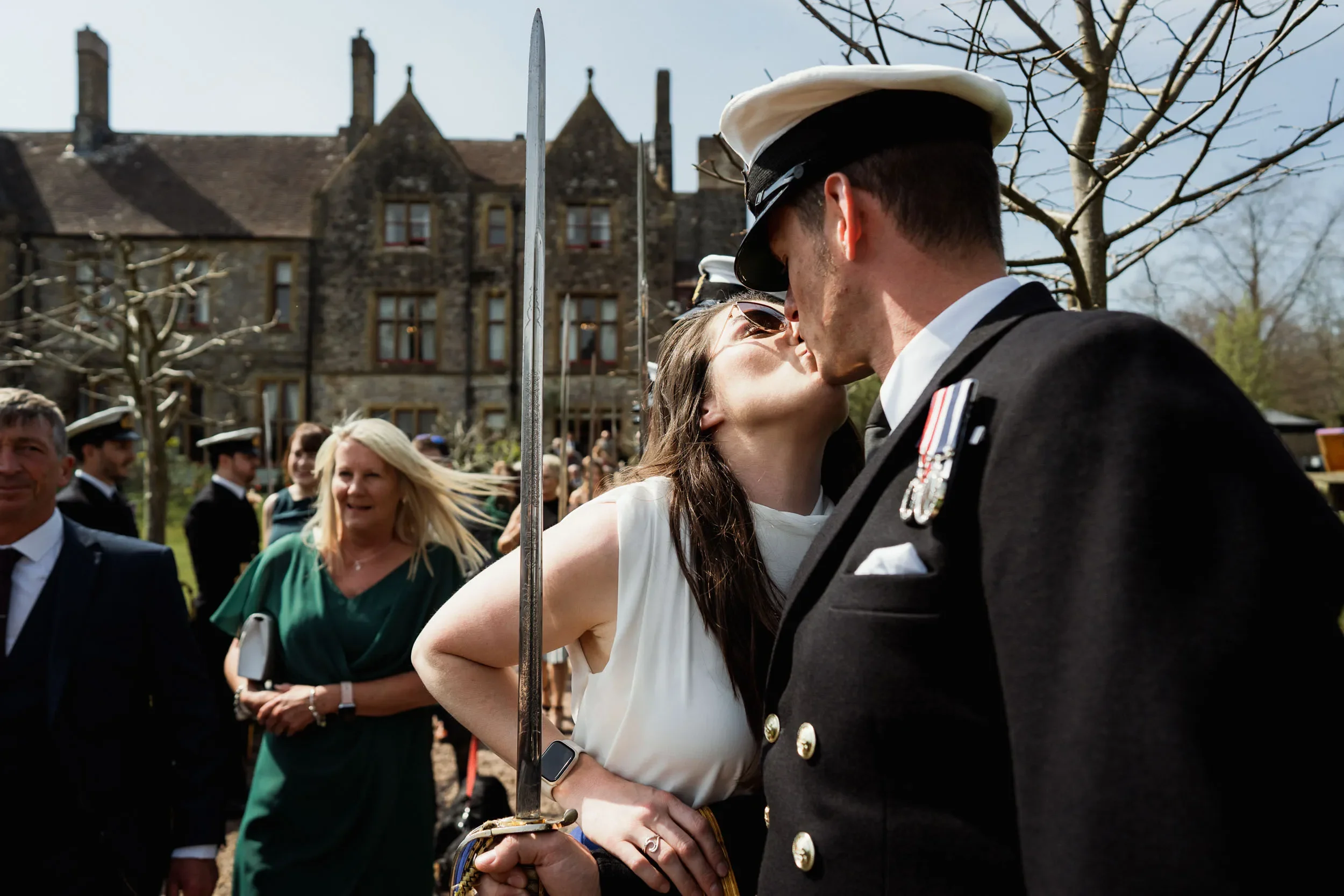 A woman and a man in military uniform kissing outdoors during a celebration, with other people in the background near a historic building and trees.