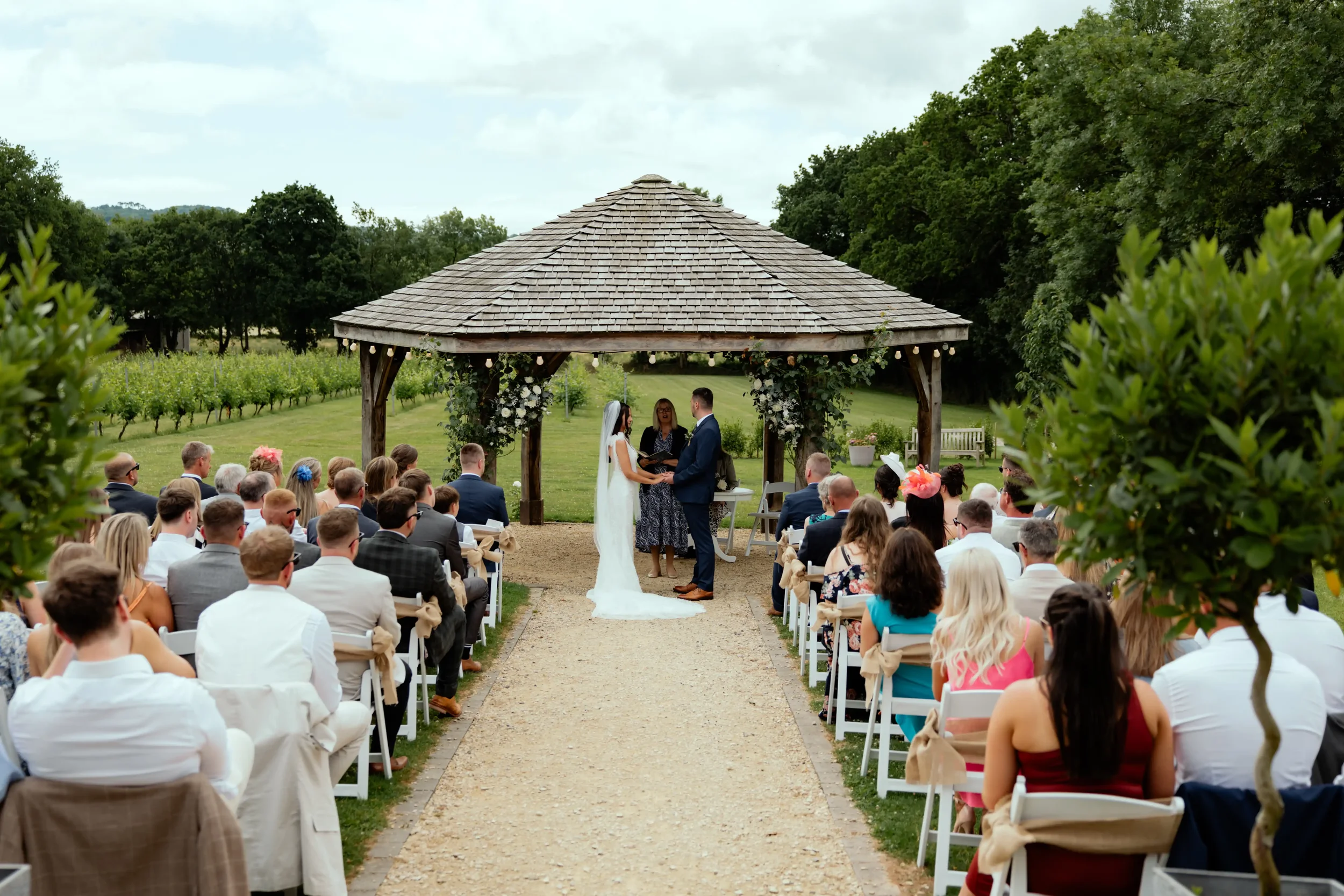 Outdoor wedding ceremony under a pavilion with a bride and groom exchanging vows, surrounded by guests seated on both sides on a gravel pathway, with green trees and fields in the background.