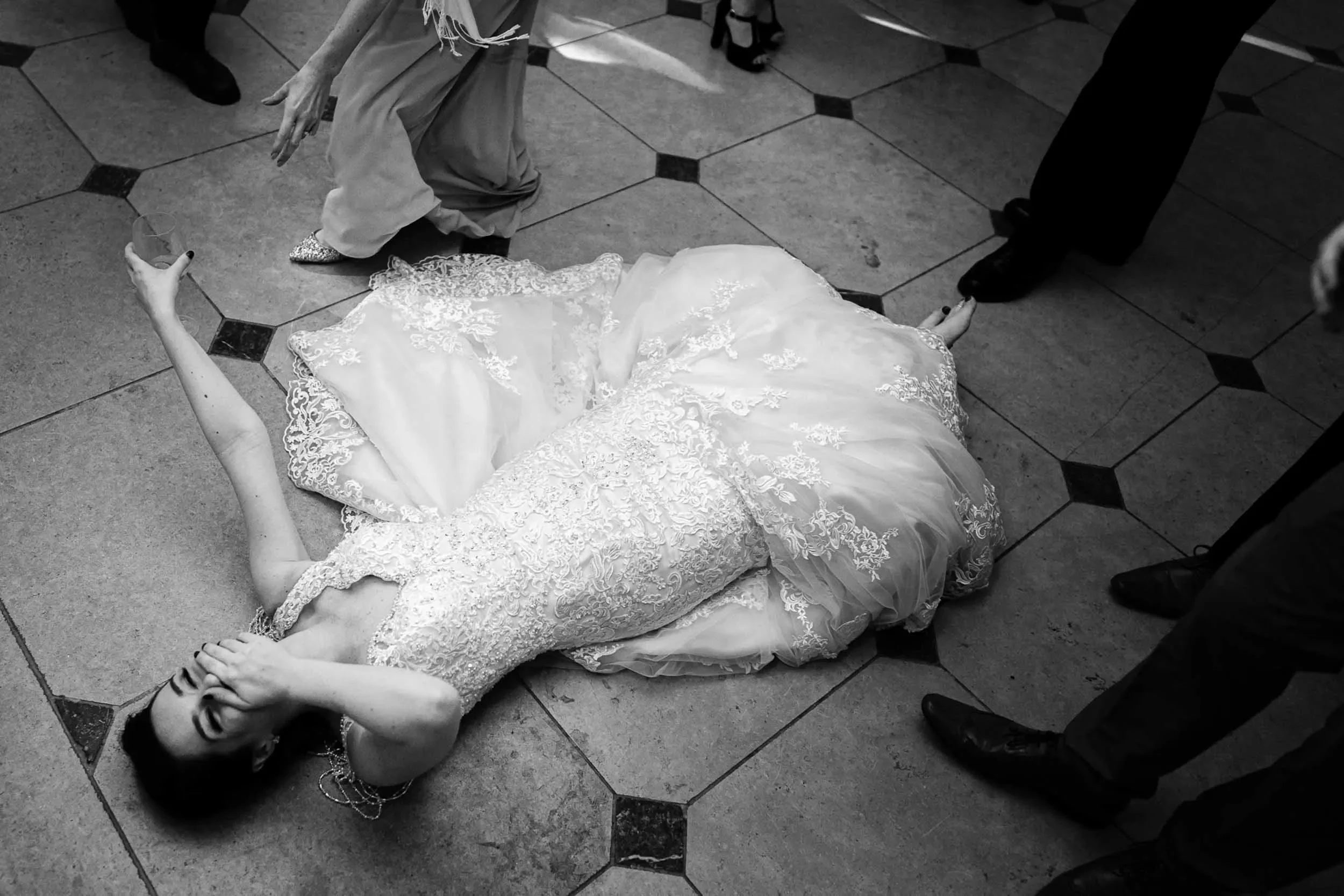 A woman in a wedding dress is lying on the floor at a wedding reception, covering her face with one hand, surrounded by guests whose legs are visible around her.