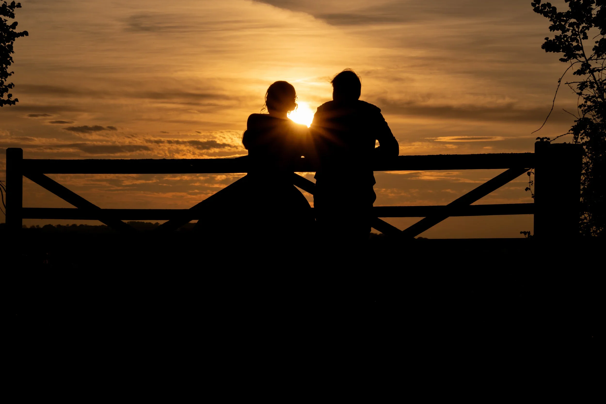 Silhouettes of a man and woman sitting together on a bench at sunset, with the sun behind them and a cloudy sky.