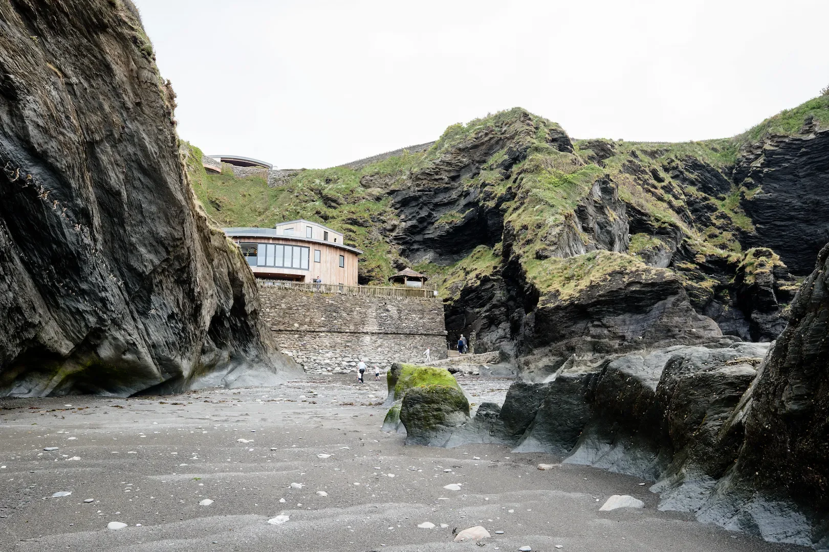 View of a rocky coastal area with a sandy beach, large rocks, and steep cliffs covered with grass. There are a few people walking on the beach near the rocks, and modern buildings are situated on the cliff side.