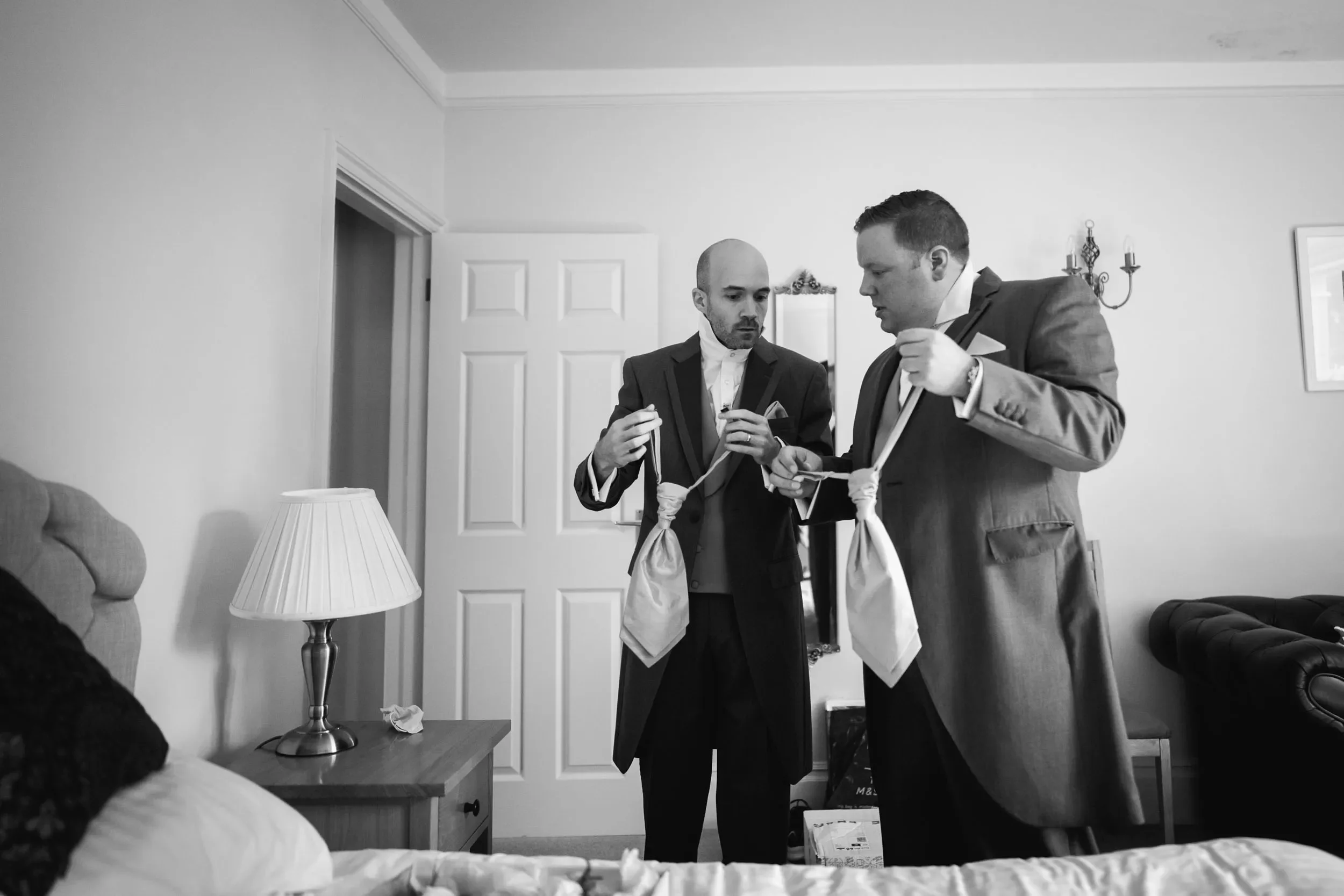 Two men in suits tying their ties in a bedroom with a bed, a nightstand with a lamp, and a wall with a mirror and candles.