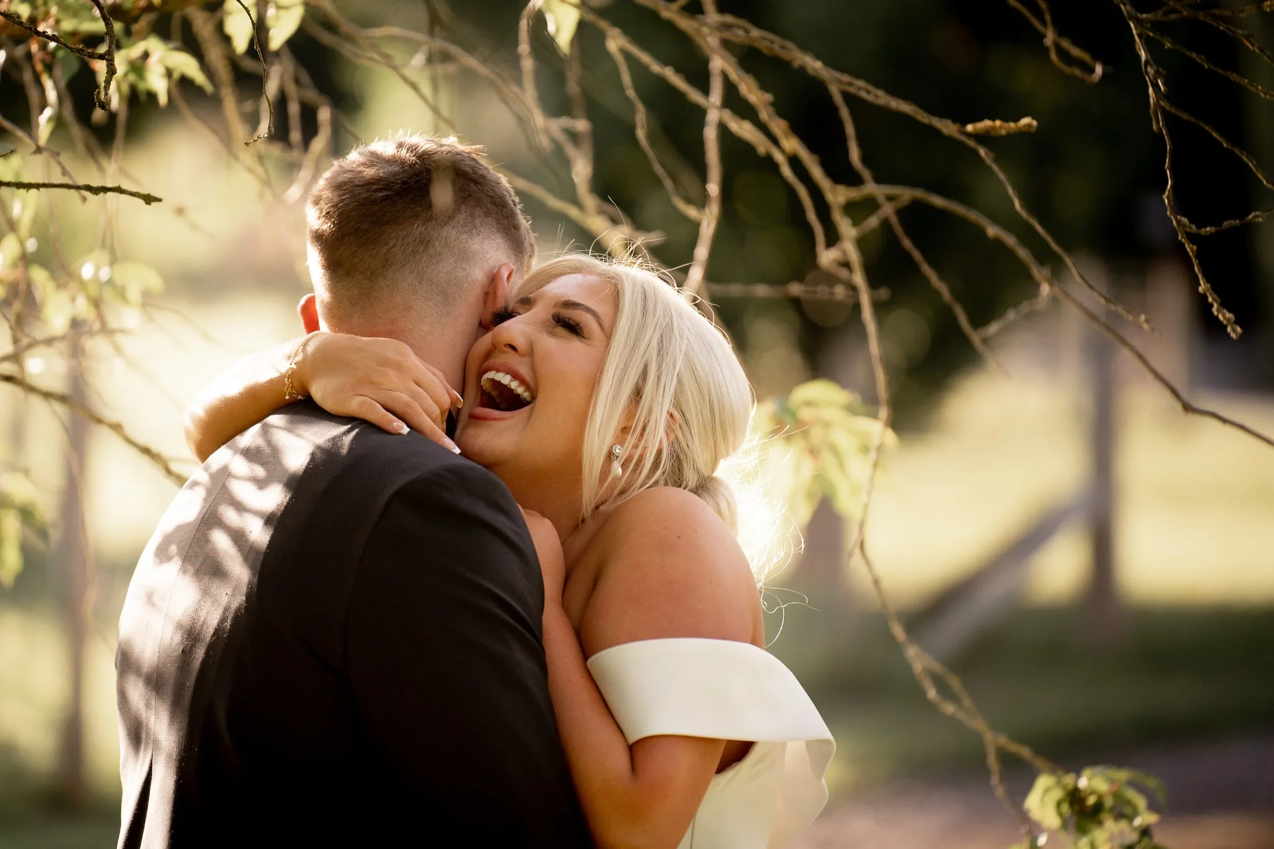 A couple sharing a joyful embrace outdoors, woman smiling with eyes closed, man with back to camera, surrounded by tree branches and sunlight.