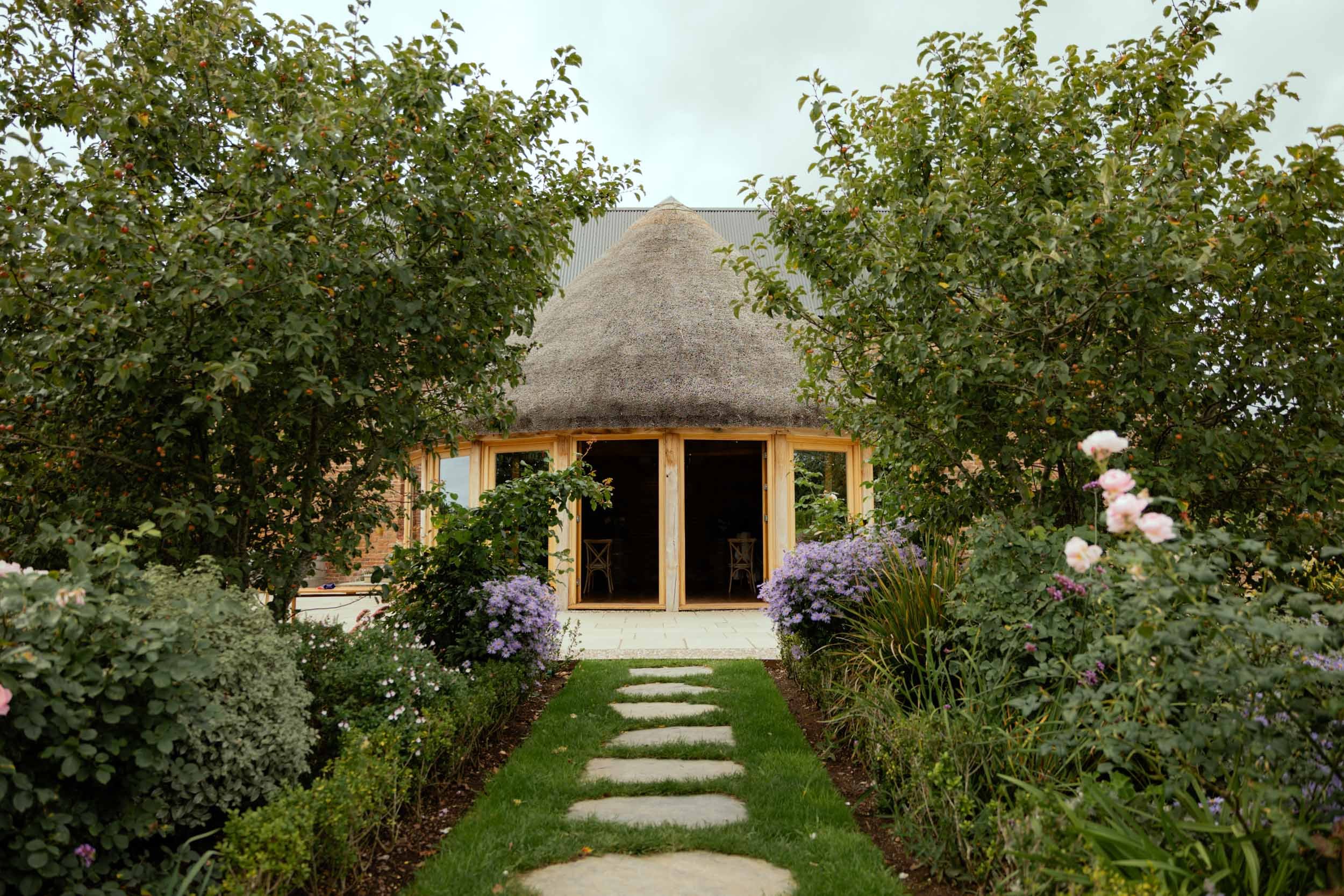Pathway of flat stones leading to a round thatched-roof house surrounded by lush greenery and colorful flowers.