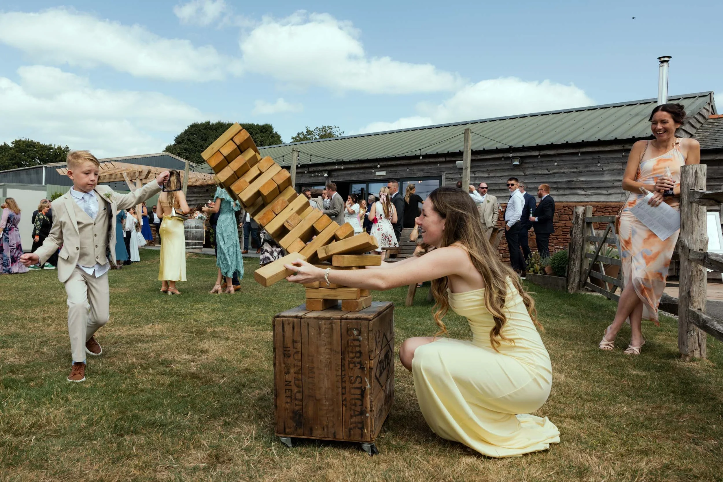 A woman in a yellow dress kneels on the grass, playfully knocking over a large Jenga game with a wooden block. A young boy in a beige suit appears to be participating in the game, reaching out toward the Jenga tower. In the background, there are seve
