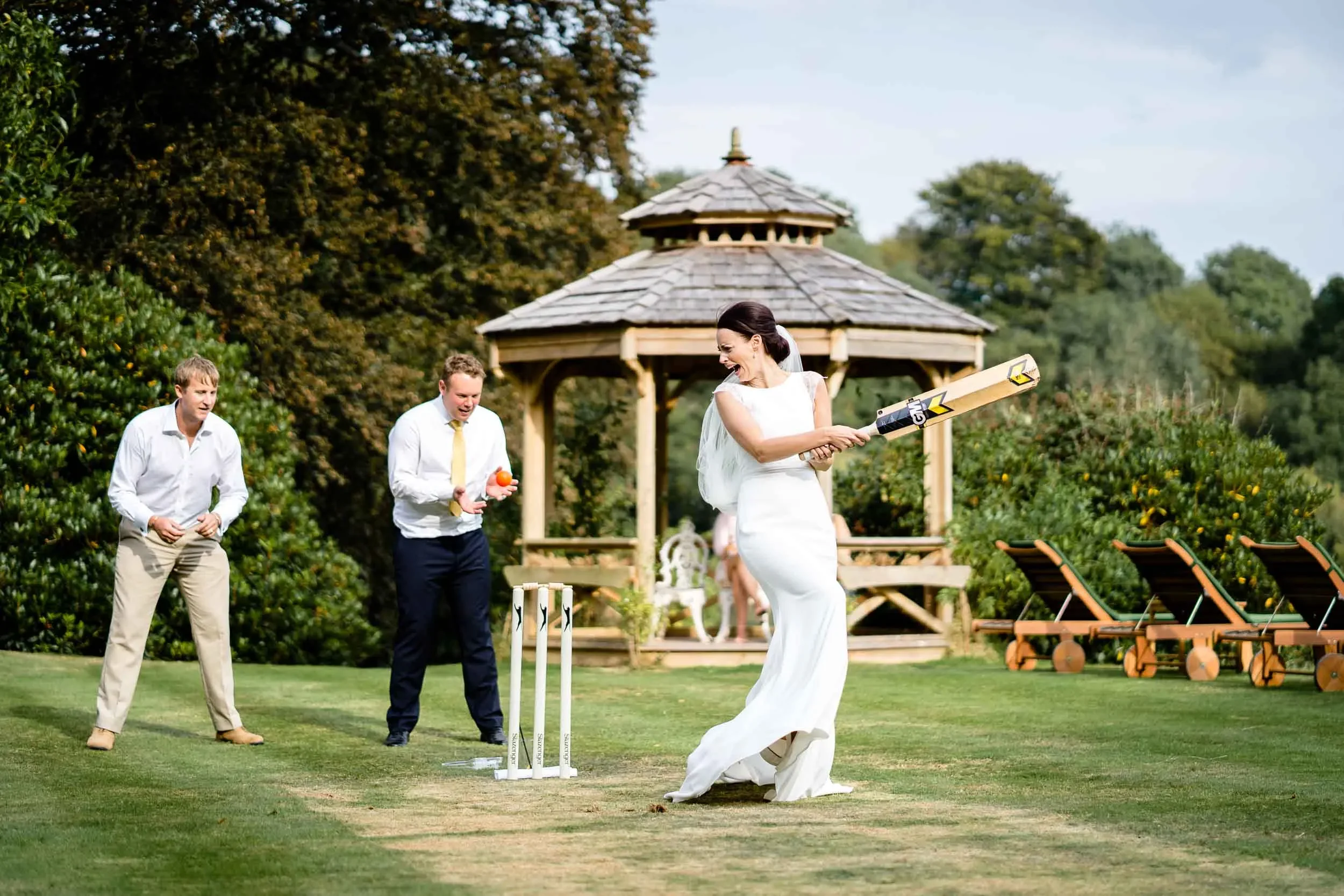A bride in a wedding dress wielding a cricket bat, celebrating on a cricket pitch with two men, one holding a ball and the other watching, in a garden setting with a wooden gazebo in the background.