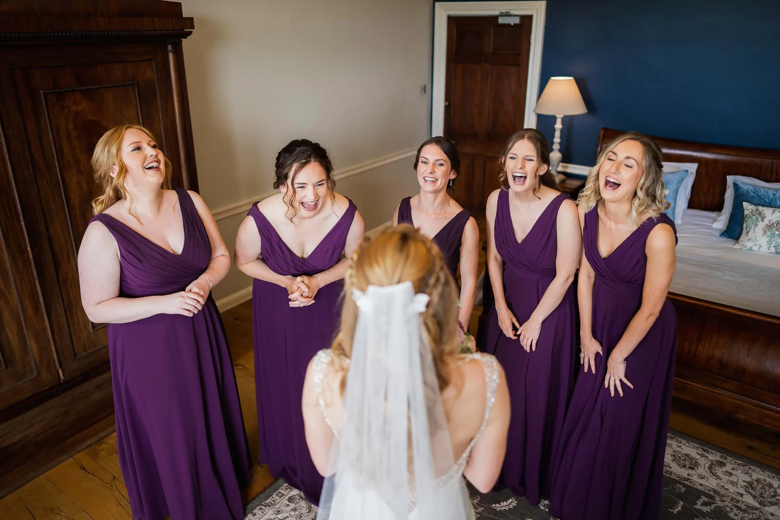 A bride in a wedding dress talking to five bridesmaids in purple dresses in a hotel room, all laughing and smiling.