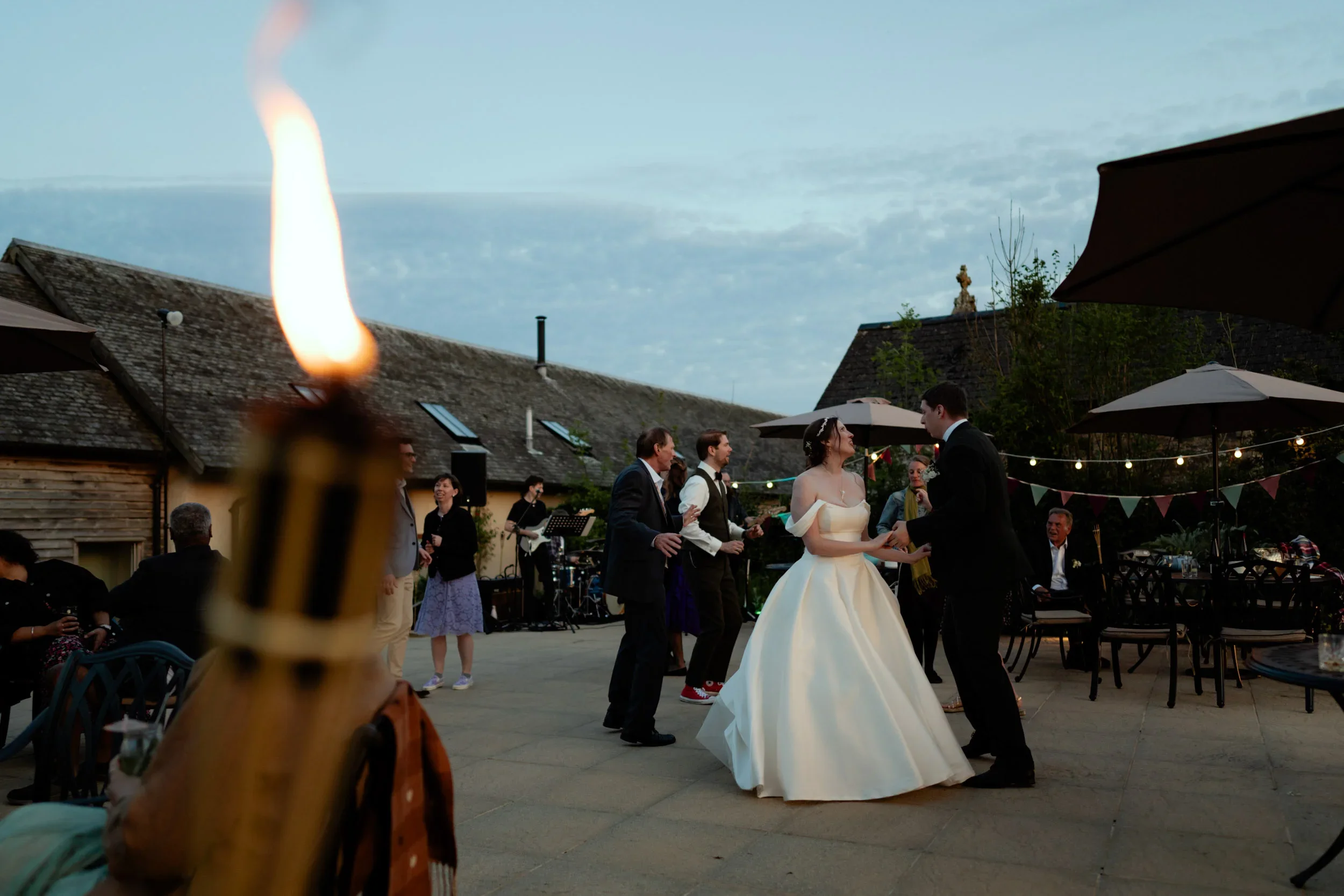 A wedding reception outdoors during the evening, with a bride and groom dancing. Guests are around, some sitting and some standing, with a band playing in the background. Decorative string lights and bunting decorate the area, and umbrellas provide s