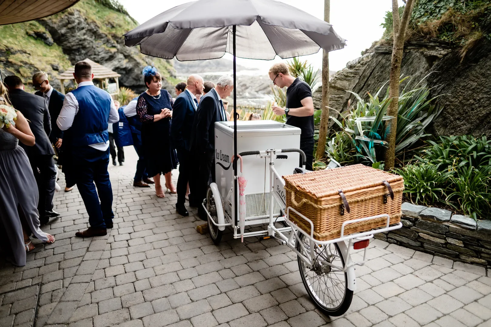 A group of people waiting in line for ice cream at an outdoor venue with rocky cliffs and lush vegetation, with a person serving from an ice cream bike under an umbrella.
