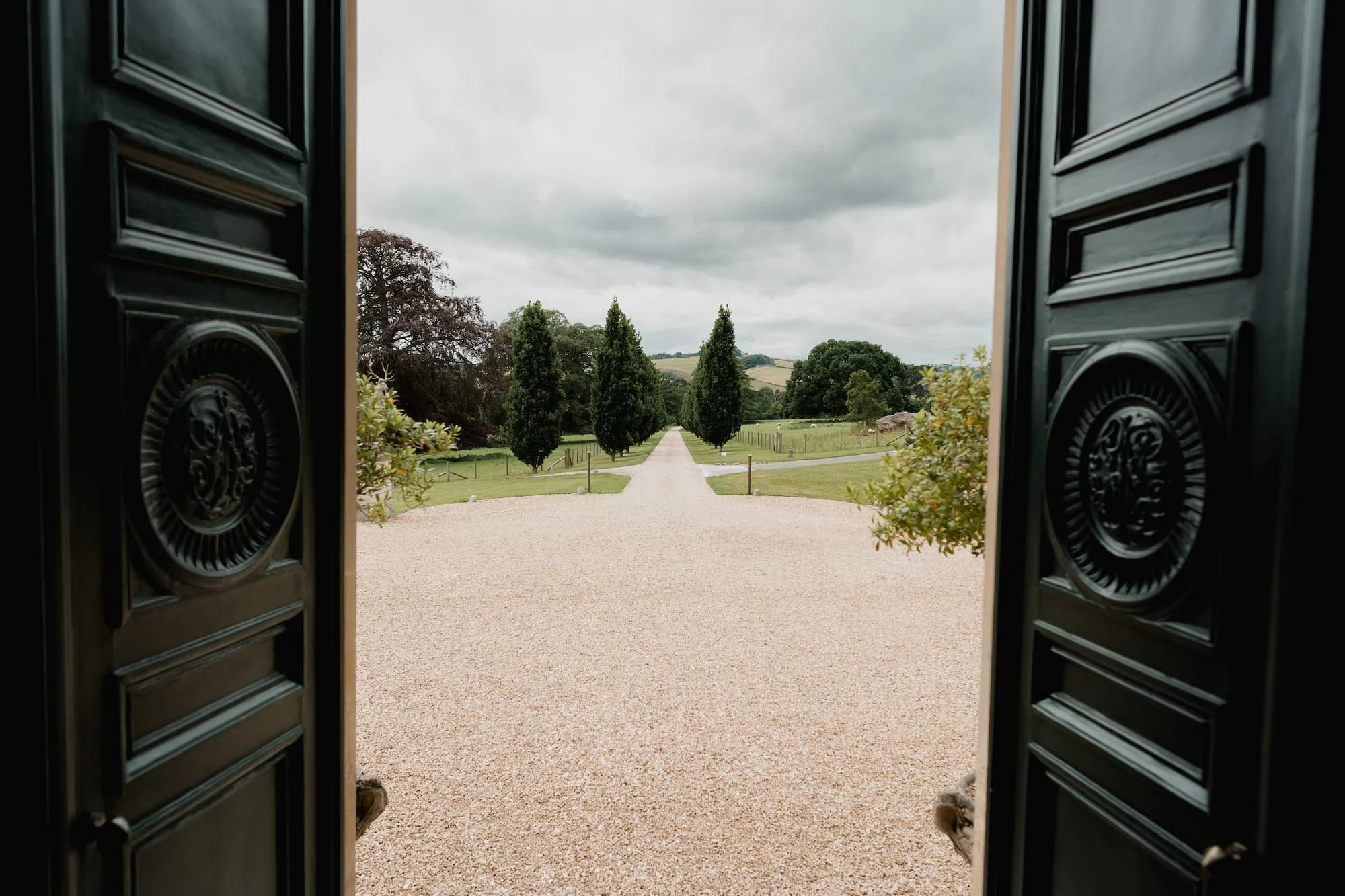 View through open black doors showing a gravel pathway lined with tall trees leading into a lush green landscape under a cloudy sky.