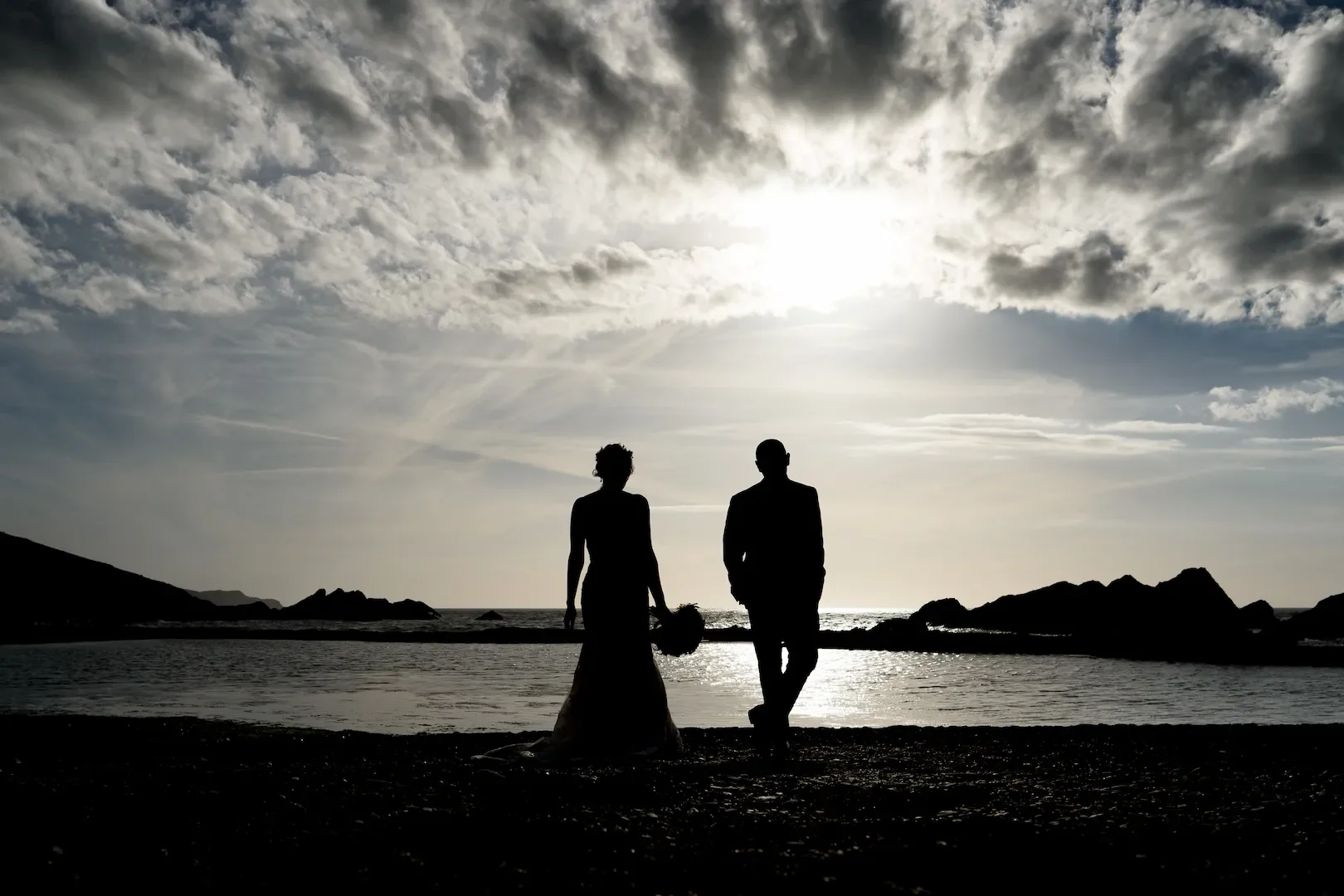 Silhouettes of a bride and groom walking on a beach at sunset with cloudy sky and rocky islands in the background.