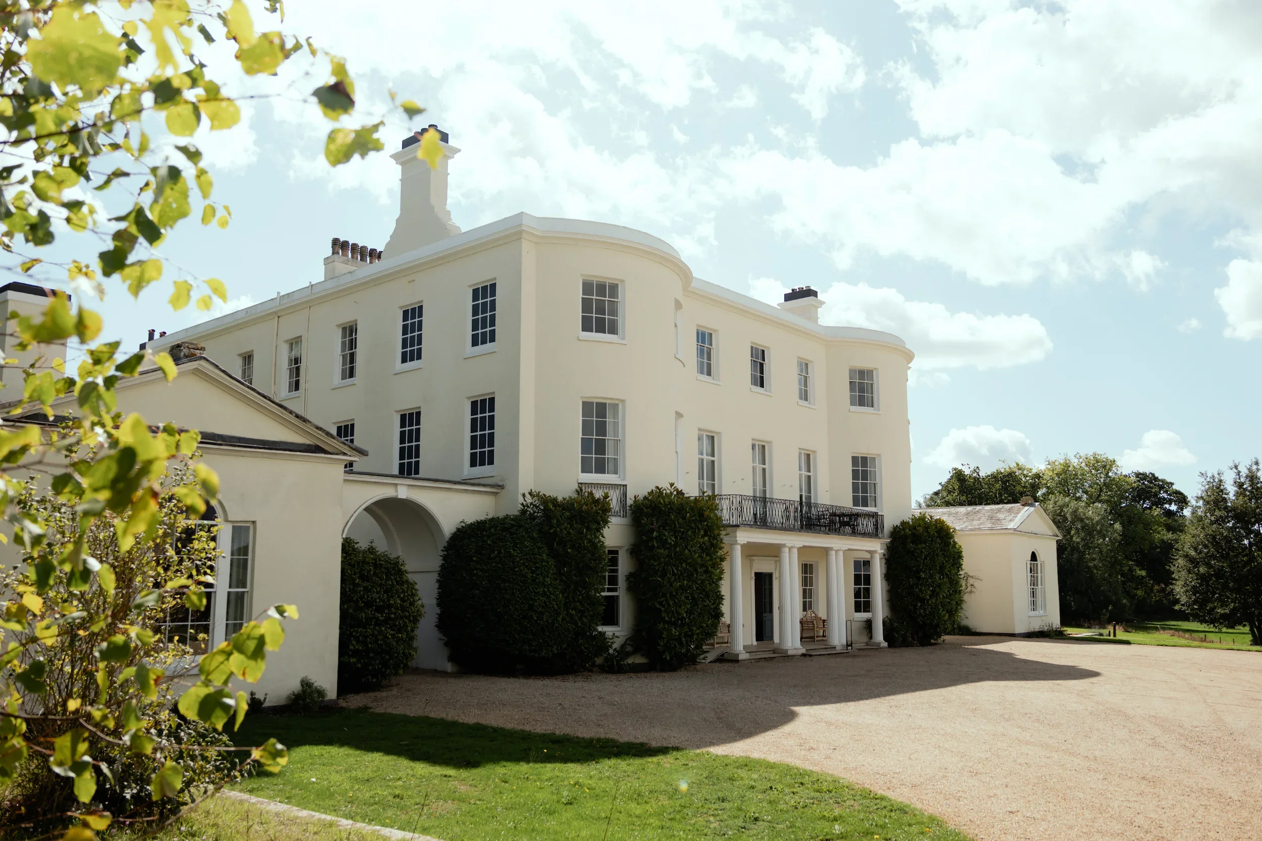 A large white historic mansion with multiple windows and columns, surrounded by greenery and a gravel driveway under a partly cloudy sky.