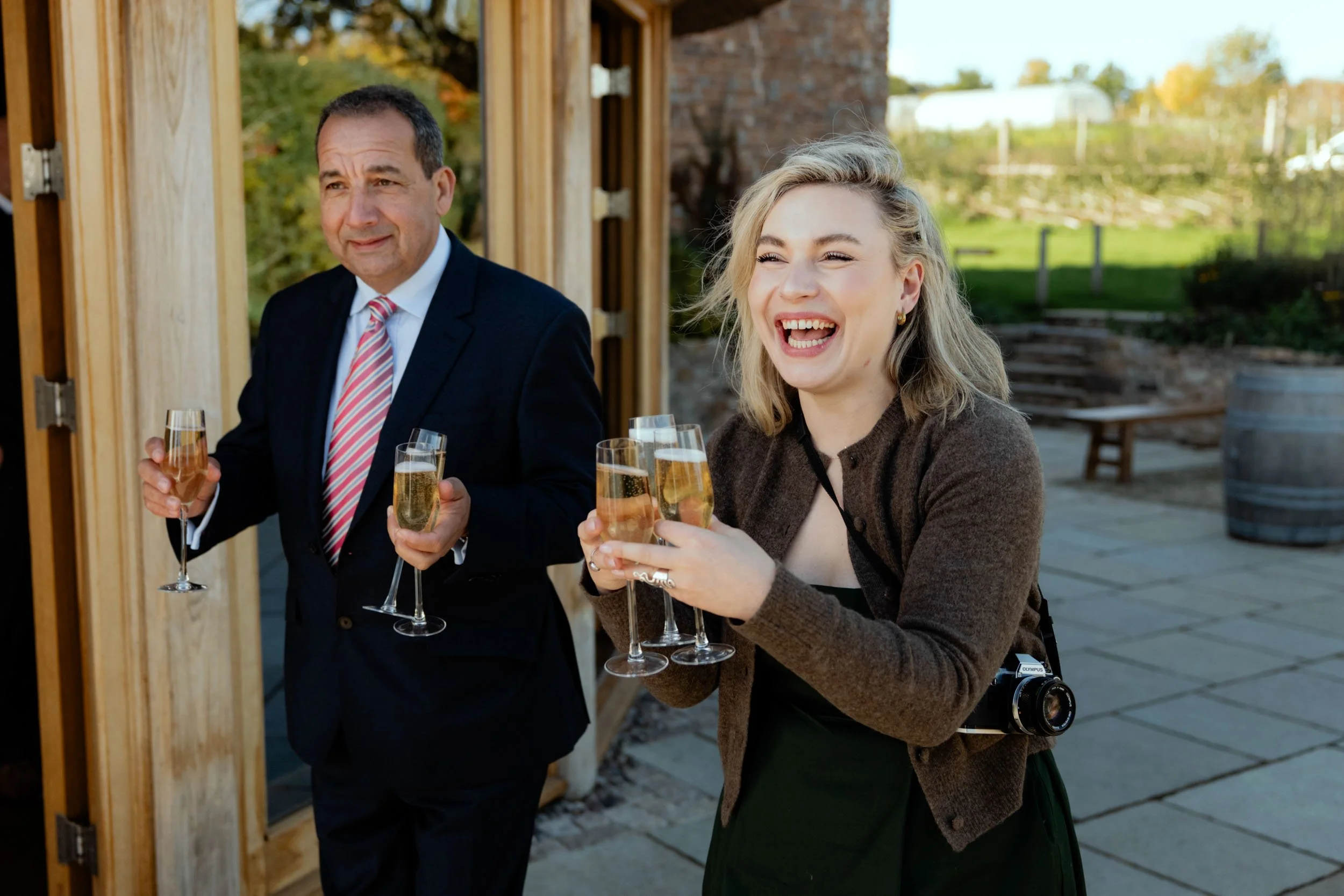 Two people holding glasses of champagne, standing outdoors with greenery in the background, one smiling and the other looking serious.