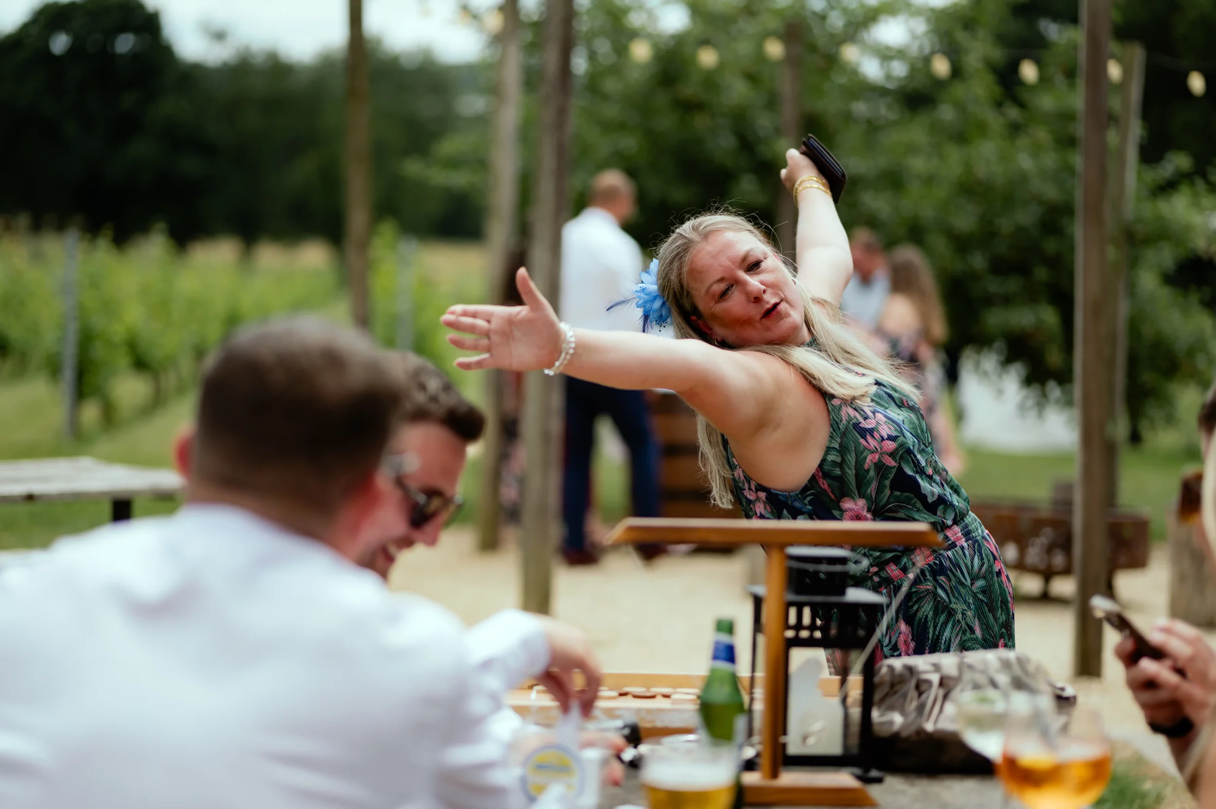 A woman dancing outdoors at a gathering, with people around her sitting at a table with drinks, in a green, wooded setting.