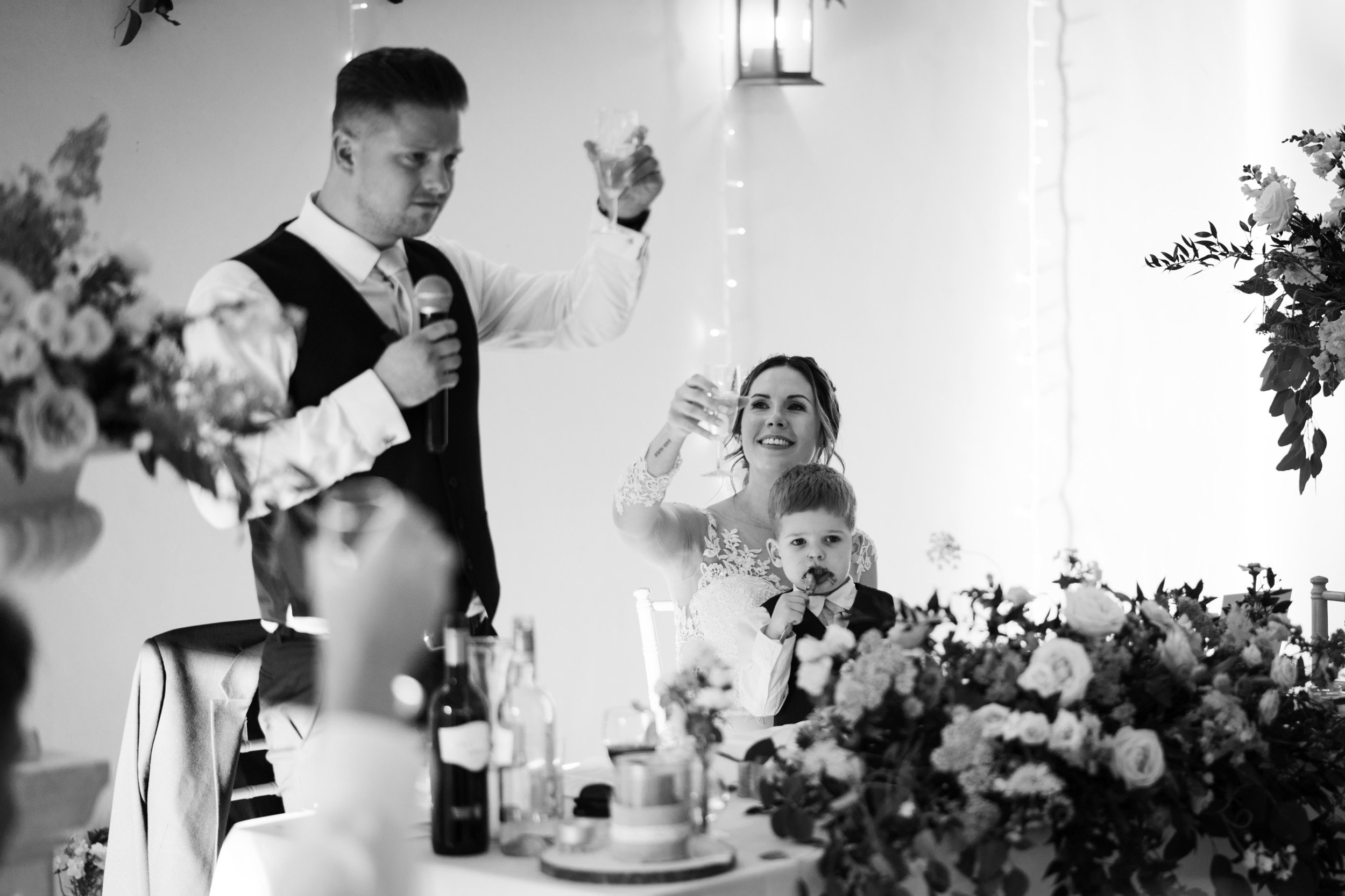 A black and white photo of a wedding reception. A man is giving a toast while holding a microphone and a glass. A woman, seated at a table adorned with flowers and a cake, smiles and raises her glass in a toast. A young boy sits on her lap, eating a 