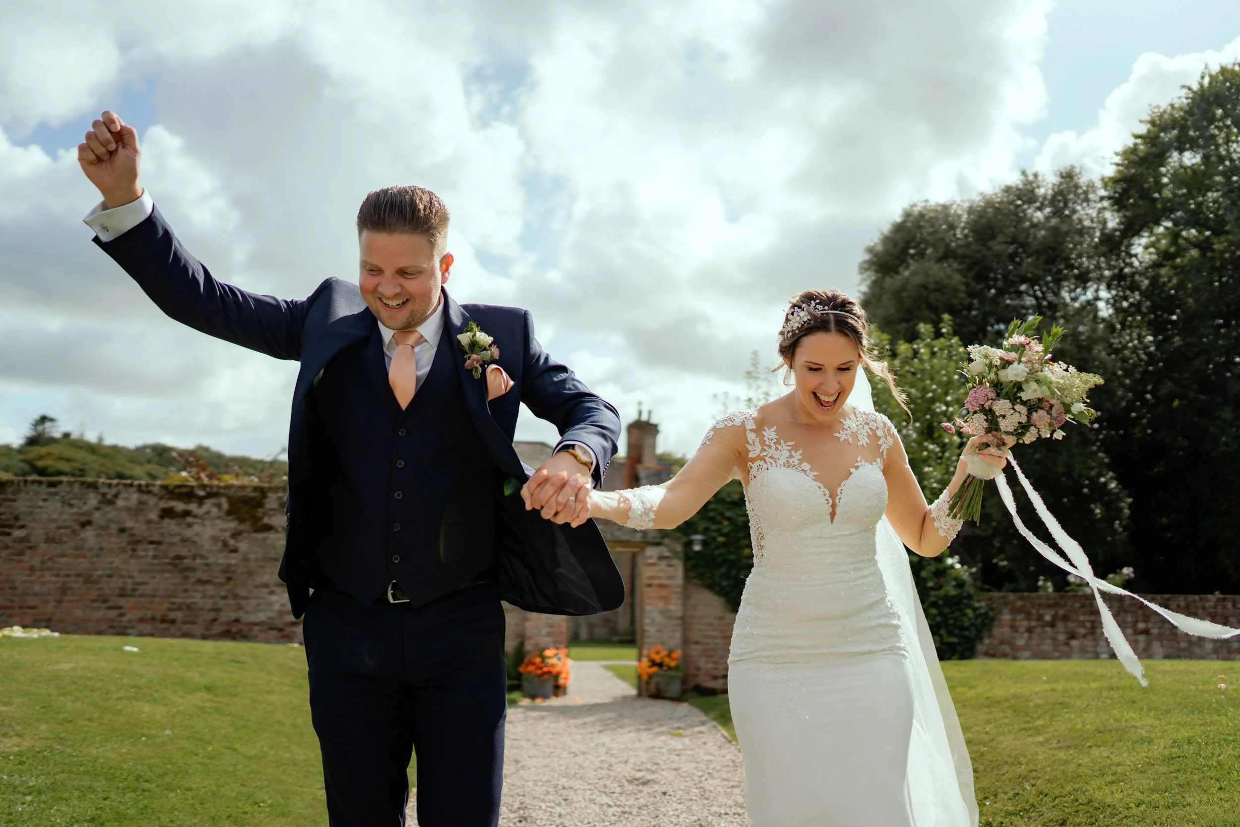 A newlywed couple, a man and a woman, celebrating outdoors on their wedding day. The man is in a navy blue suit with a pink tie and boutonniere, raising his arm in joy. The woman is in a white lace wedding gown, holding a bouquet of flowers, and smil
