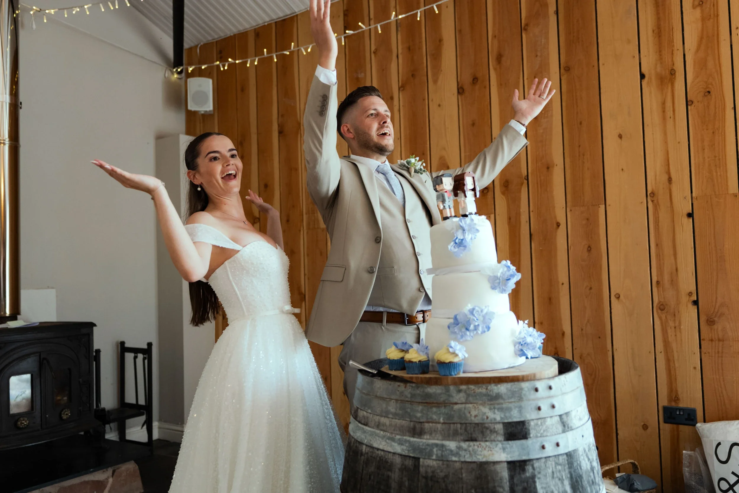 A bride and groom celebrating their wedding with a cake in front of a wooden wall, with the groom raising his hands and the bride smiling with her arms raised.
