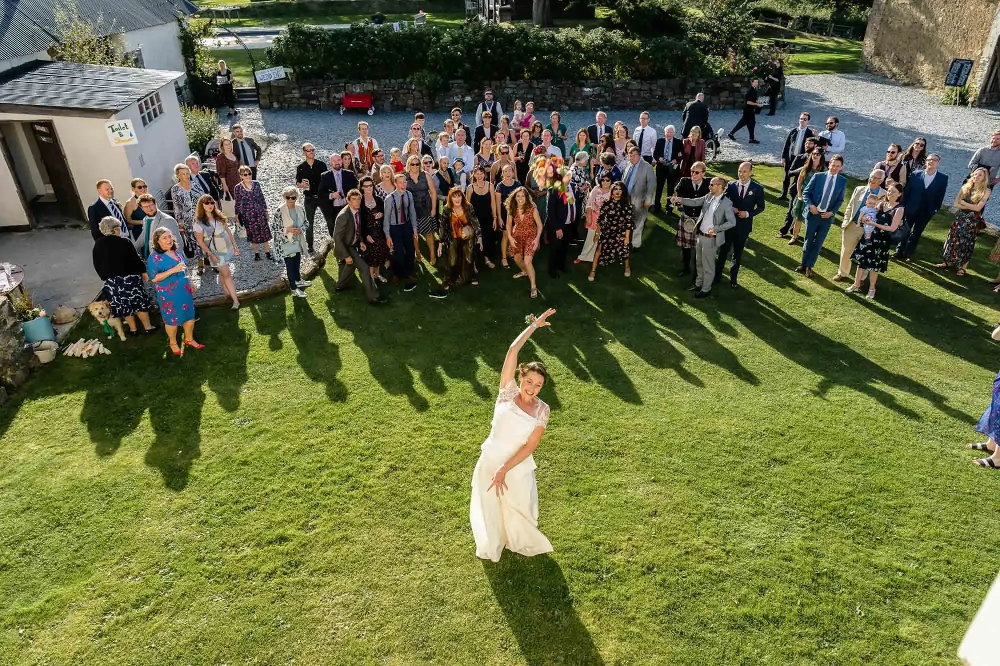 A bride in a white dress dancing alone on a grassy area at a wedding reception, surrounded by a large group of guests who are standing and watching her. The scene is outdoors with sunlight casting shadows on the grass.