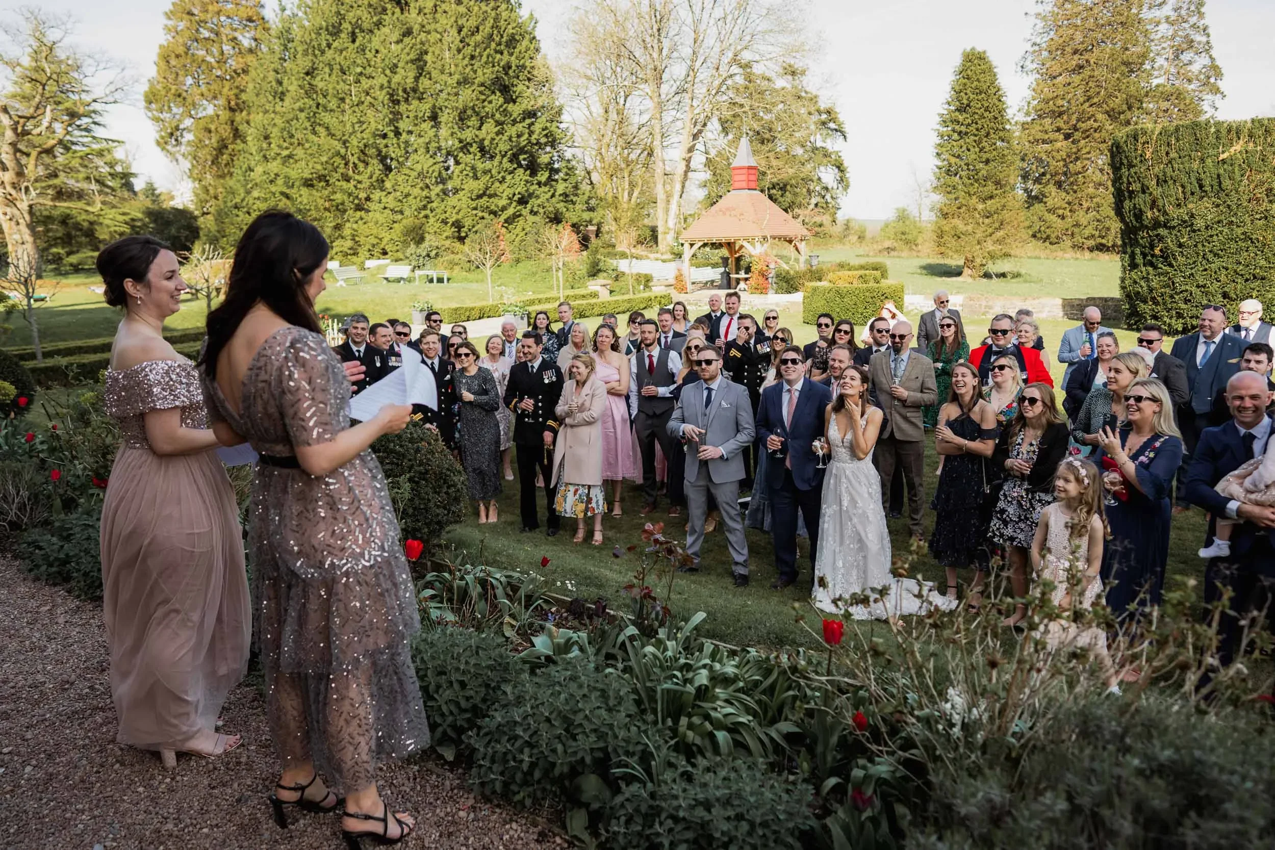 Guests at an outdoor wedding reception featuring a garden, green trees, and a small pavilion in the background, listening to two women giving a speech.