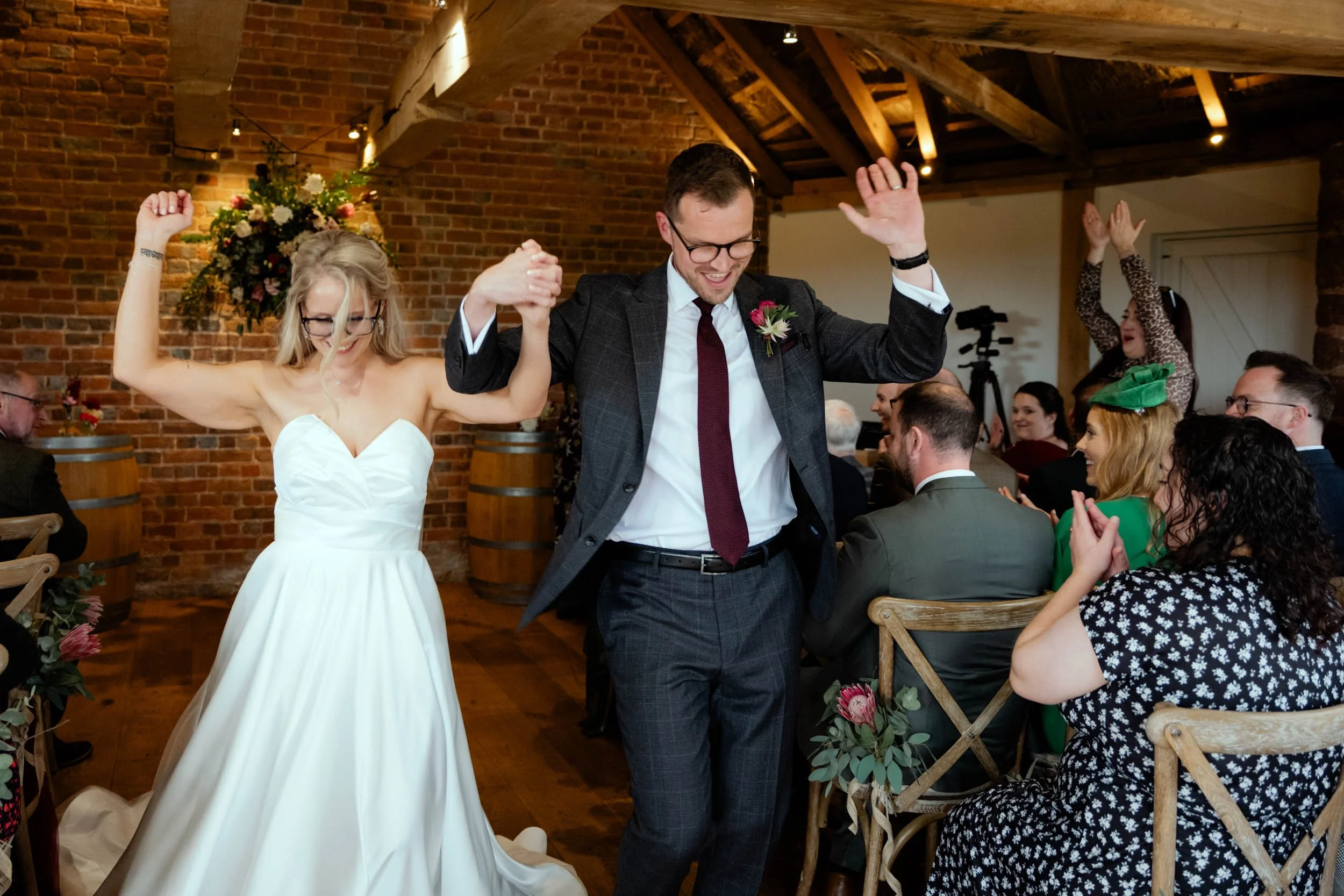 Bride and groom dancing at wedding reception with guests clapping and celebrating in a rustic venue with brick walls and wooden beams.