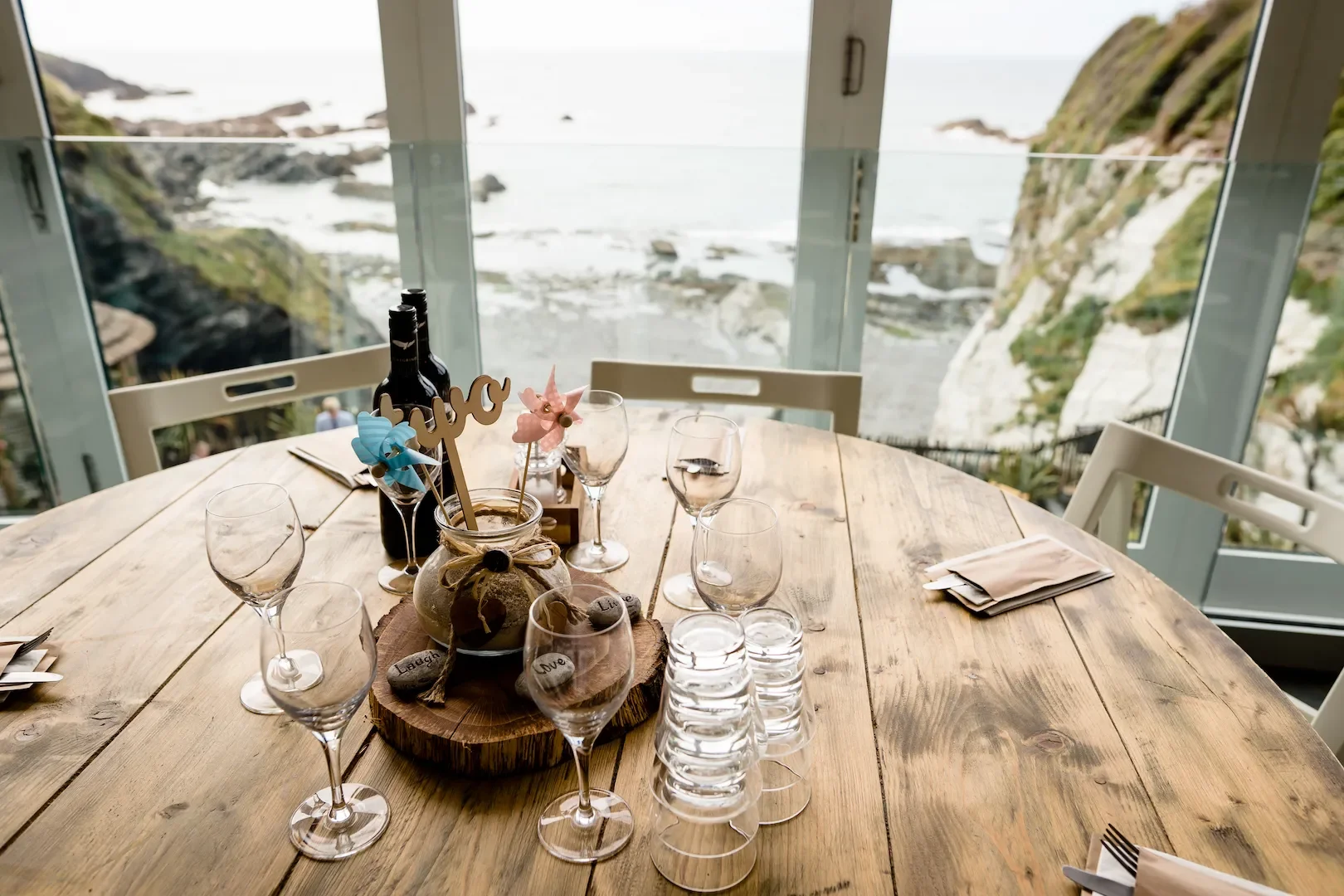Empty round wooden table set with empty wine glasses, small glasses, napkins, and a decorative centerpiece with a small jar, flowers, and a wooden cutout that says 'love'; table overlooks a coastal landscape with rocky cliffs and the sea visible thro