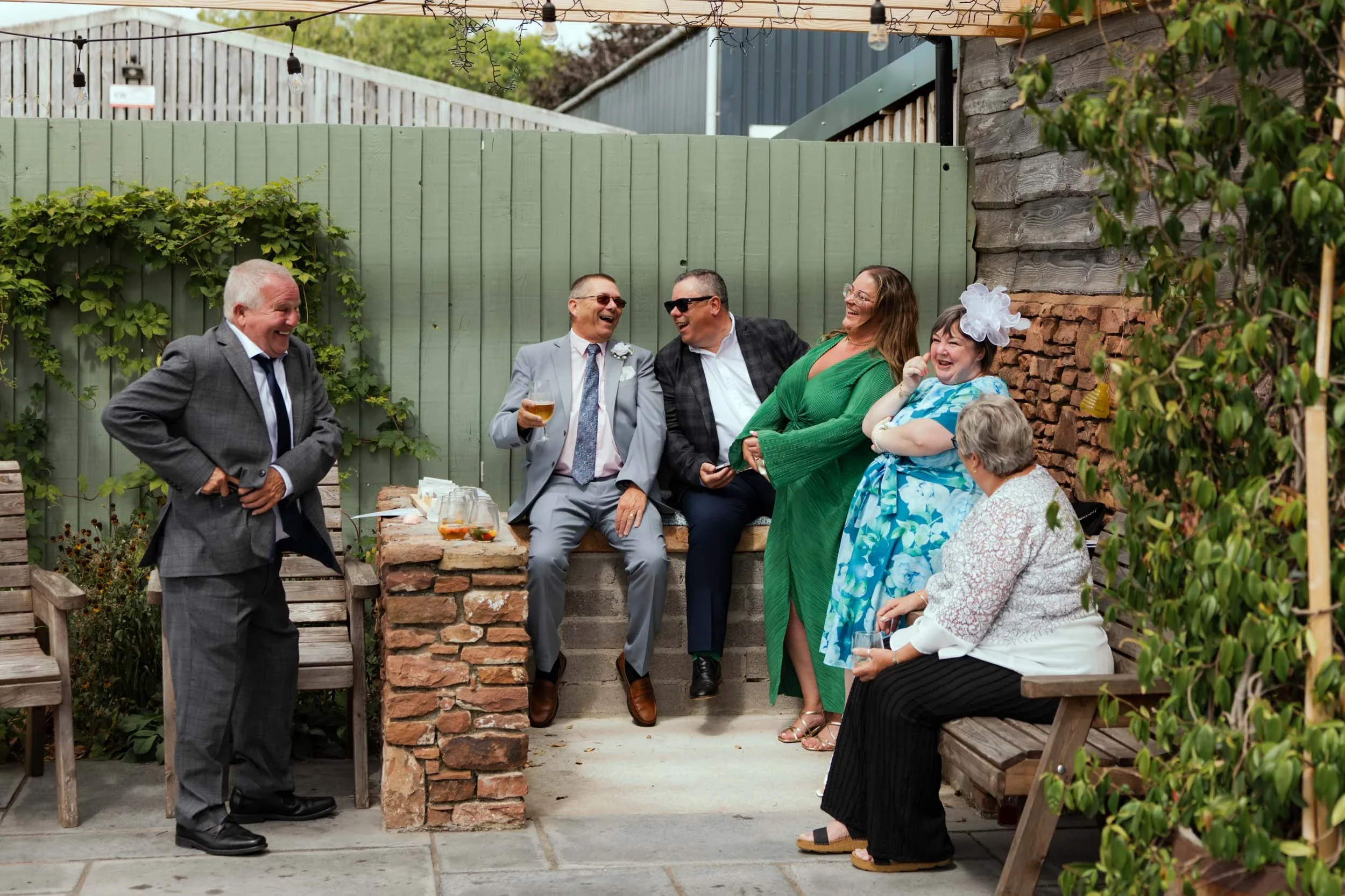 People at an outdoor gathering, laughing and talking, including women and men dressed in formal and casual attire, with some holding drinks, seated on benches and standing in a backyard with greenery, wooden fencing, and string lights overhead.
