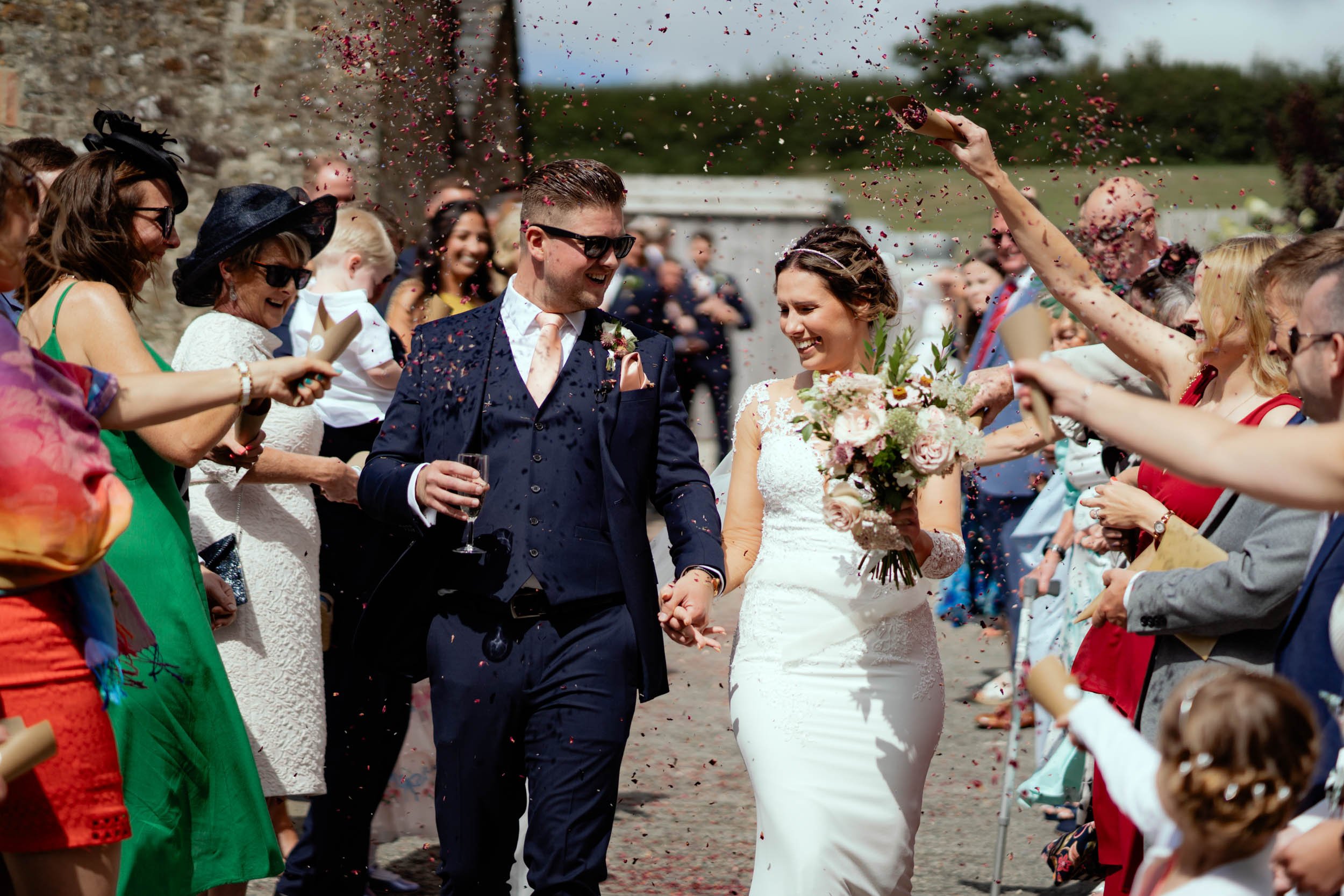 Bride and groom holding hands walk through guests throwing confetti at a wedding celebration outside on a sunny day.