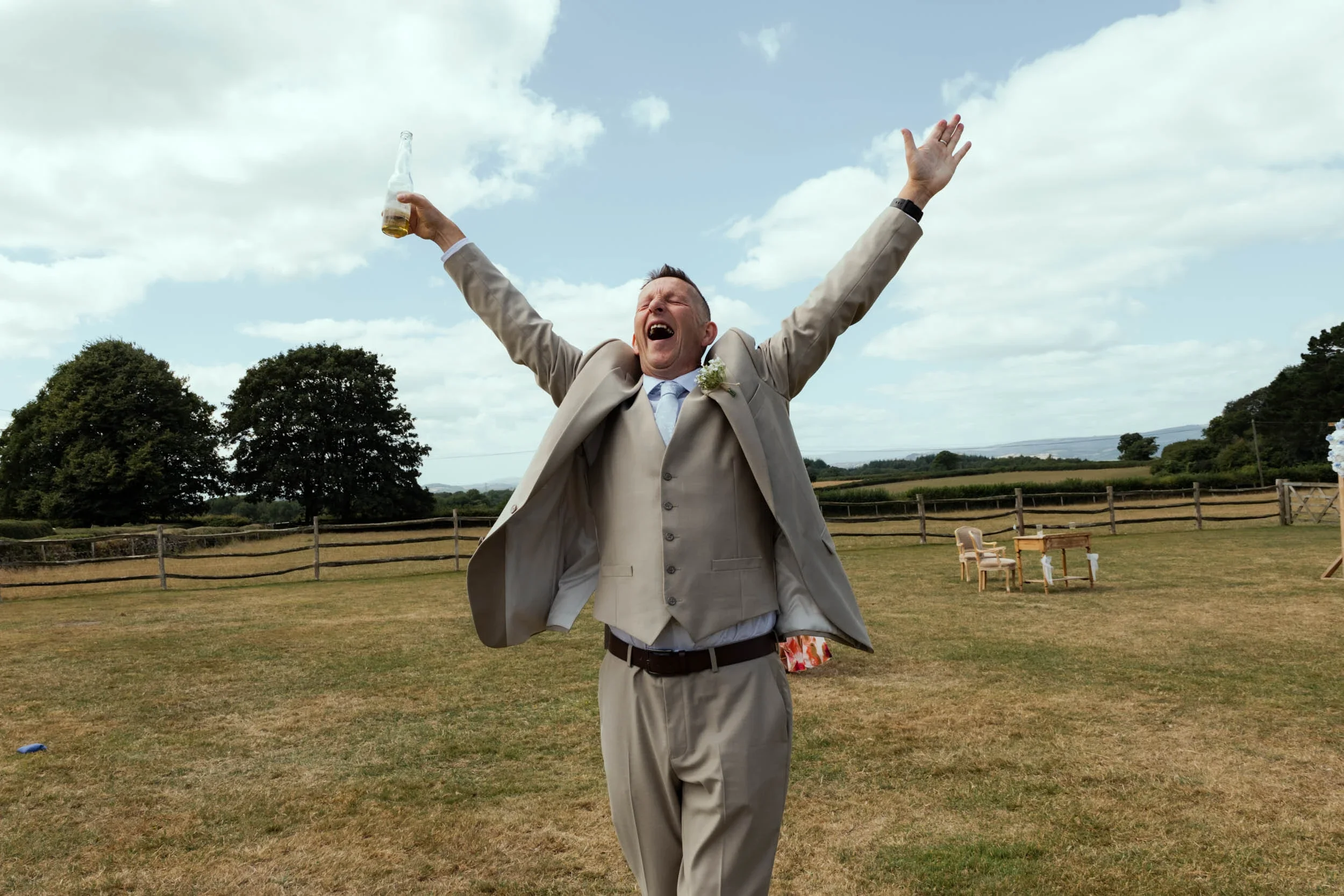 A man in a beige suit and blue tie celebrating outdoors with arms raised, holding a drink, on a grassy field with trees and scattered chairs in the background.