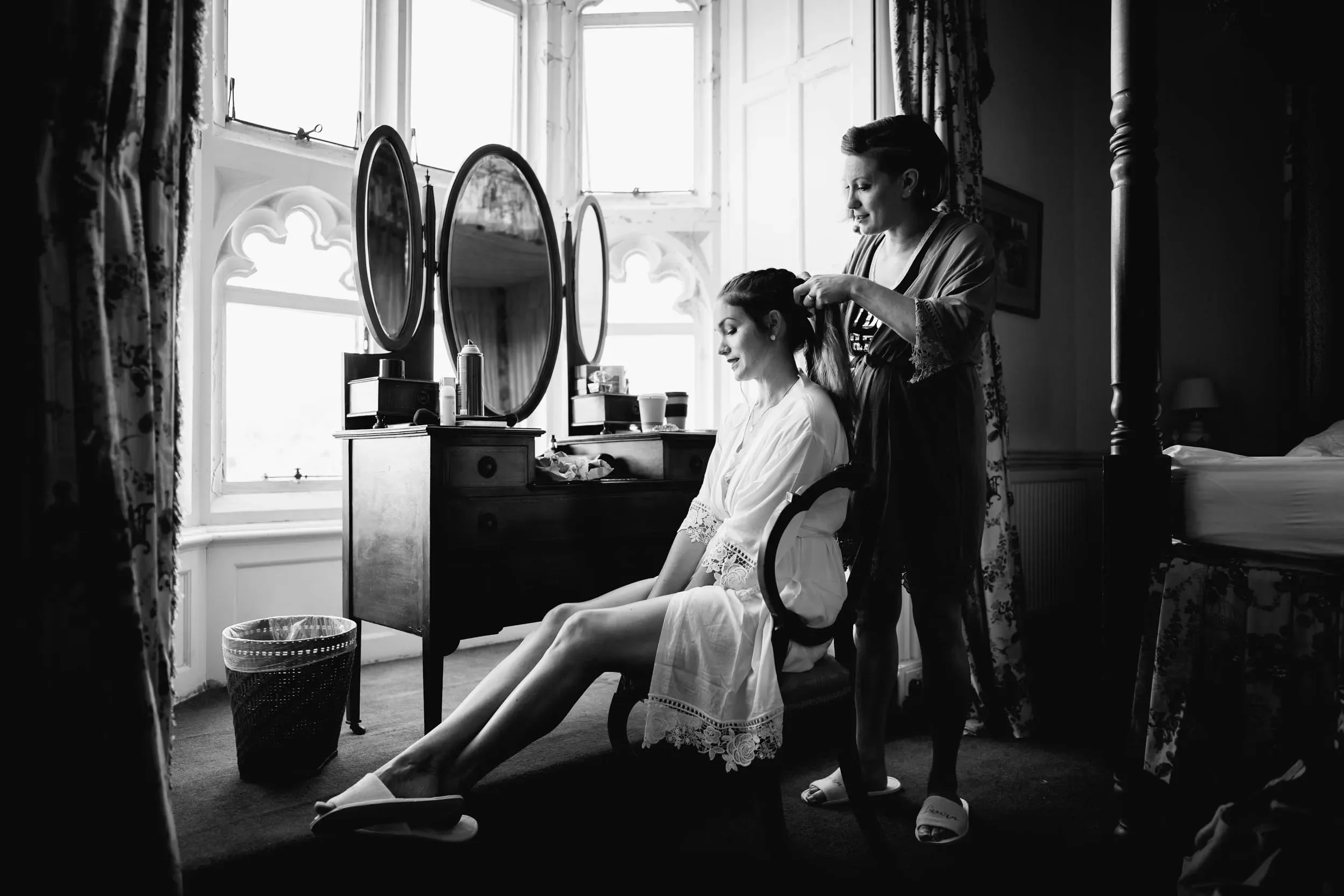 A woman is sitting in a chair, getting her hair styled by another woman in a bedroom with large windows and floral curtains.