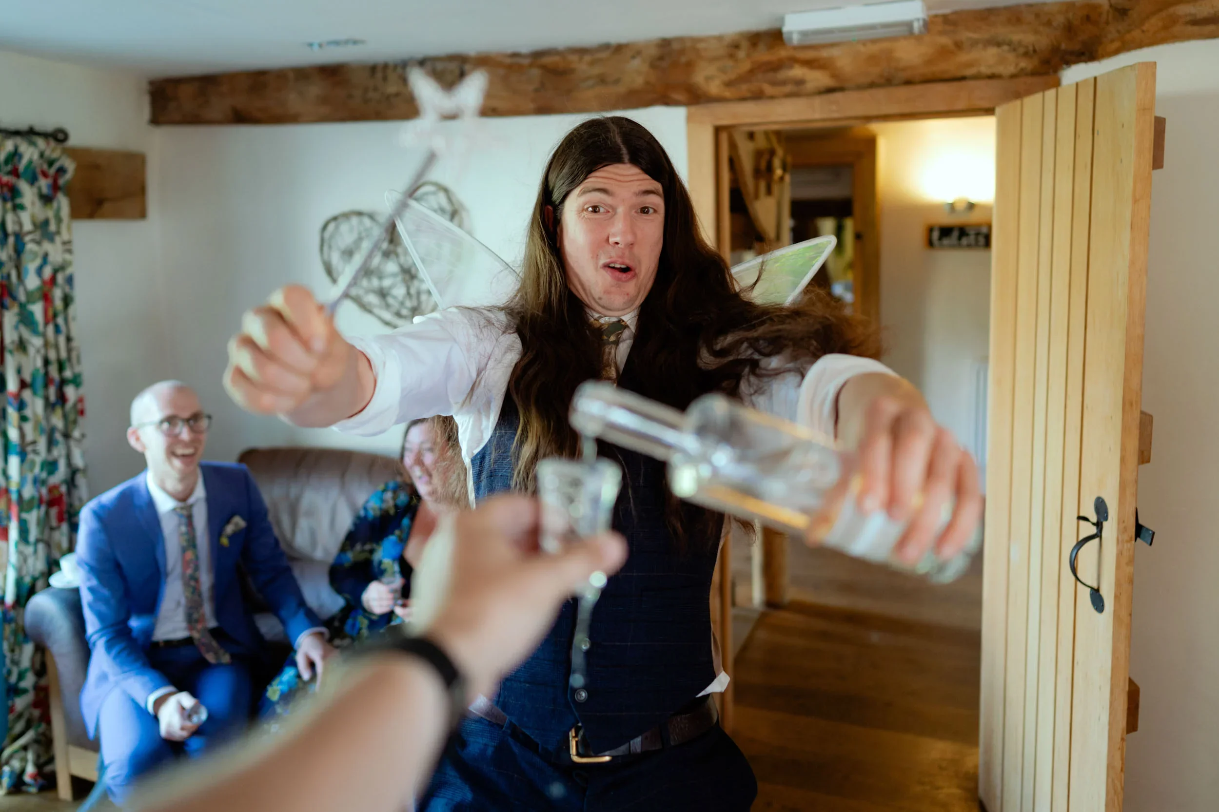 A man with long hair is reaching out with a glass bottle and a shot glass. Three people sitting on a couch behind him are laughing and smiling at his actions. The scene takes place in a cozy, rustic room.