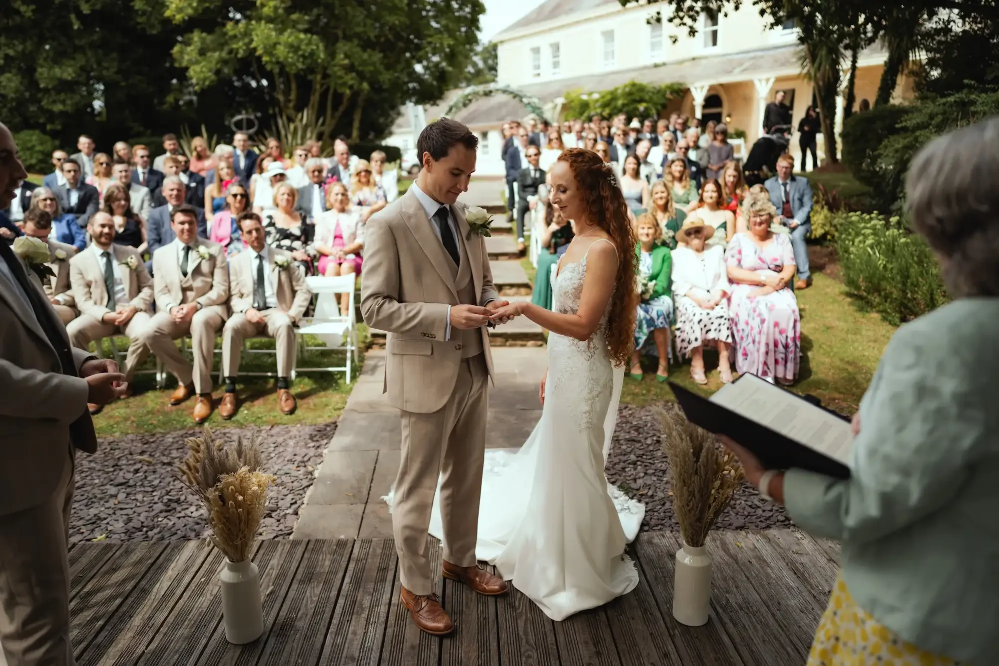 A couple exchanging vows during an outdoor wedding ceremony, with guests seated and standing behind them in a garden setting.