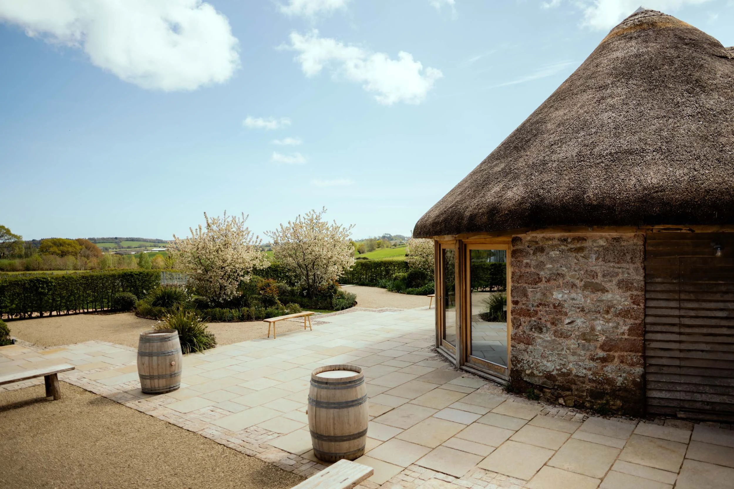 A rustic stone building with a thatched roof, glass doors, and a scenic outdoor patio with wooden barrels, benches, flowering trees, and distant green fields under a blue sky with clouds.