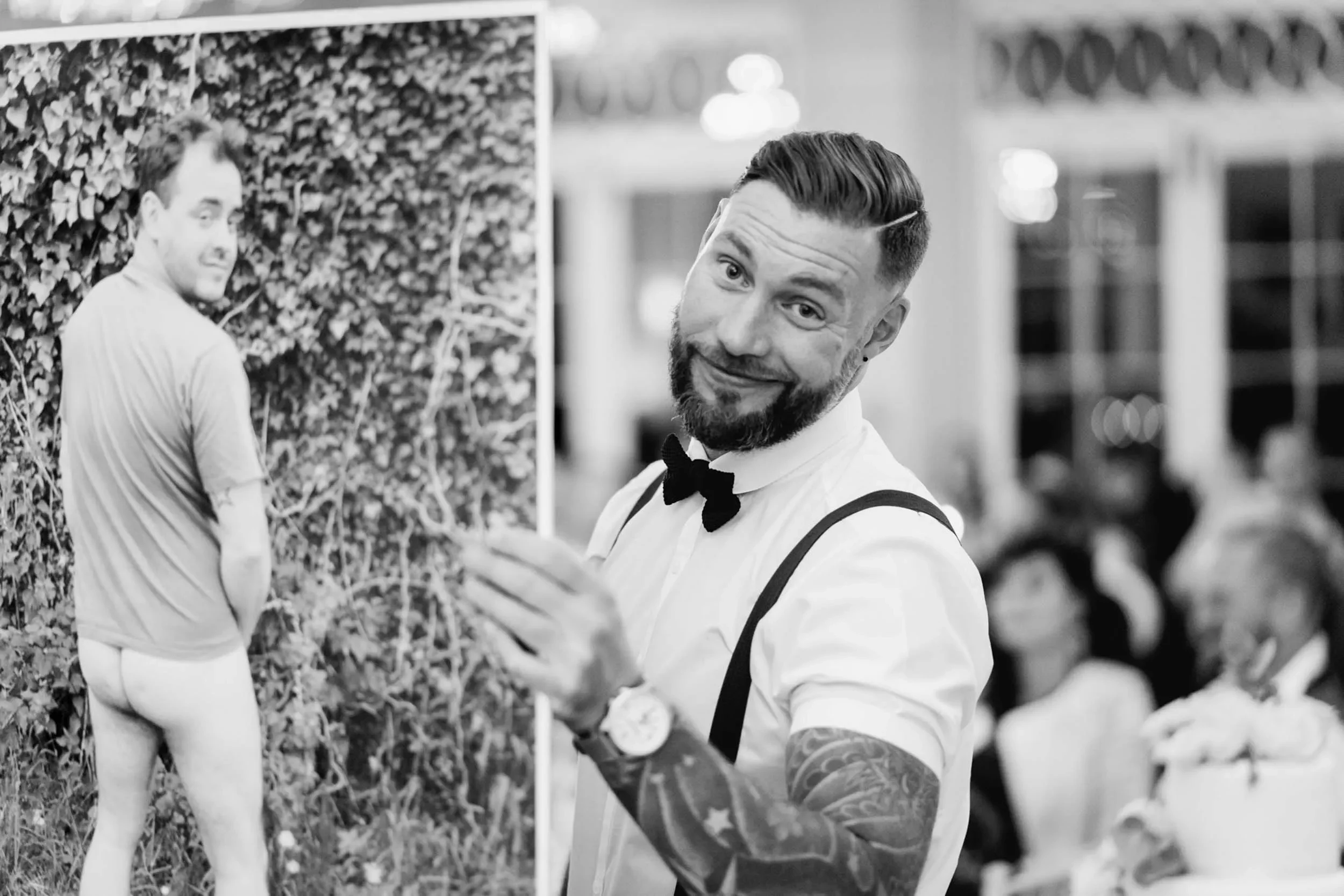 A man with a beard, wearing a bow tie, suspenders, and a watch, is smiling and pointing towards a photo of another man with a beard, wearing a T-shirt, standing outdoors against a hedge, with the back turned and looking over his shoulder.