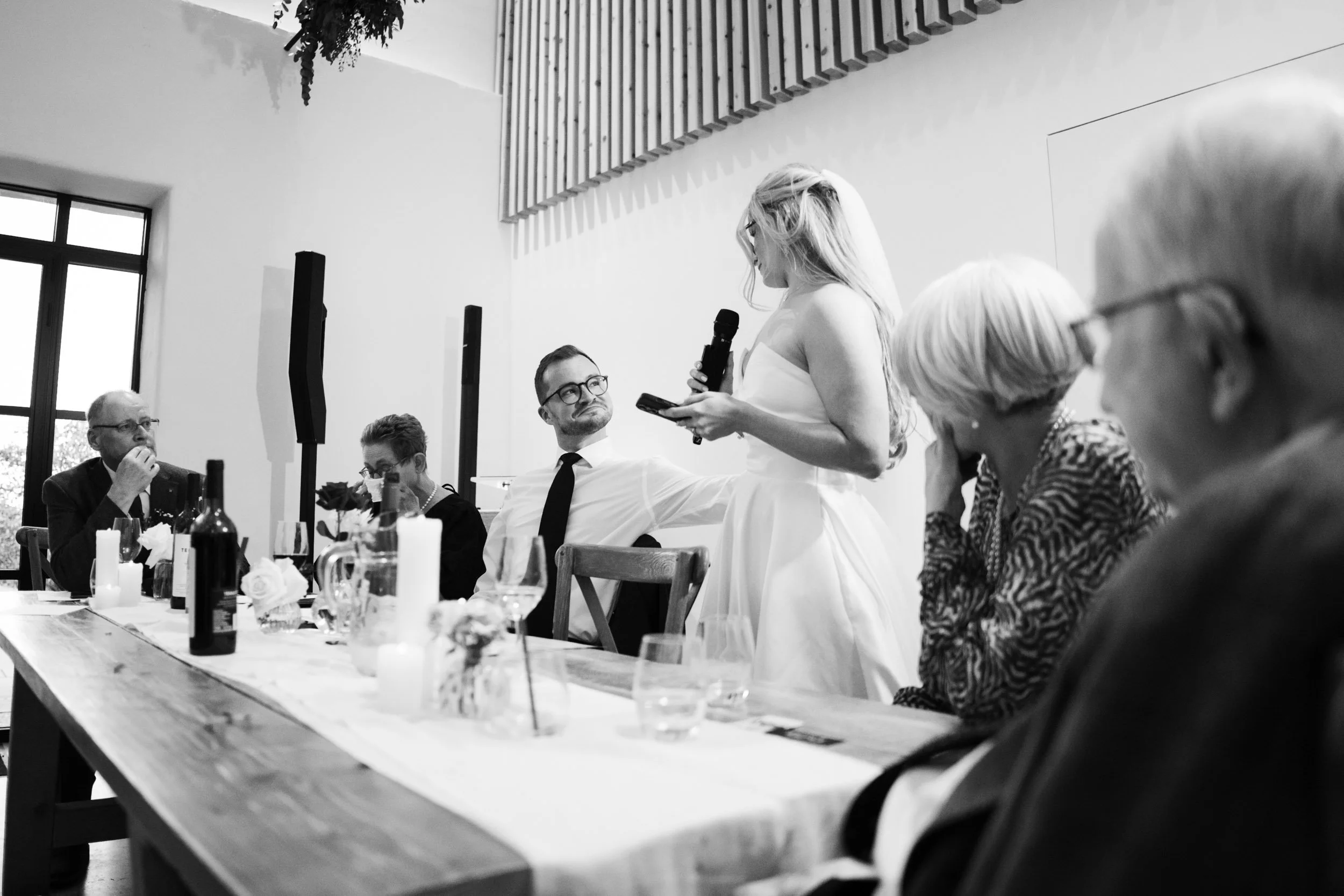 Black and white photo of a woman in a wedding dress giving speech at a table during a wedding reception, with seated guests listening.