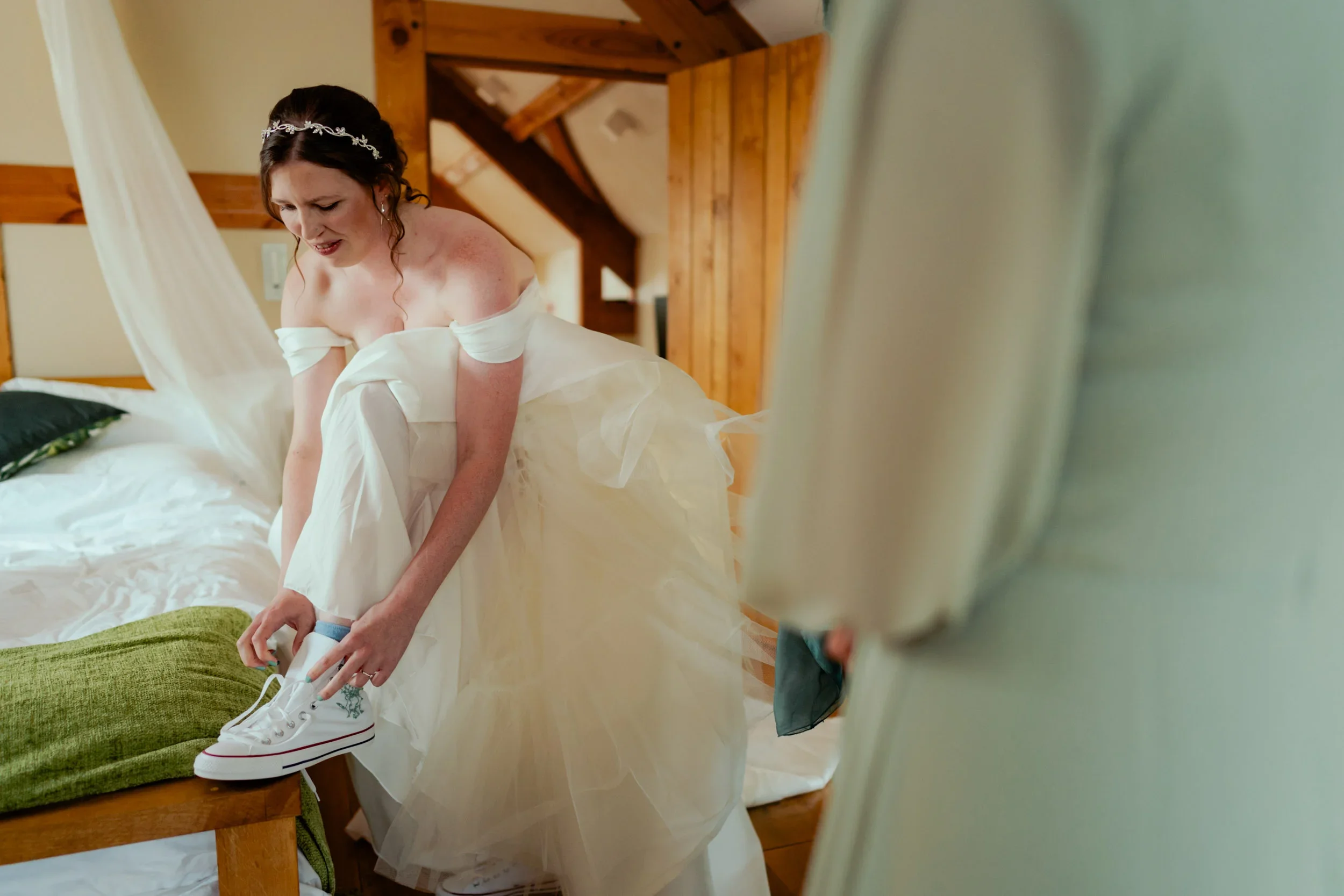 A bride in a wedding dress is putting on white sneakers on her right foot while standing on a bed in a wooden room. She looks focused and slightly emotional.
