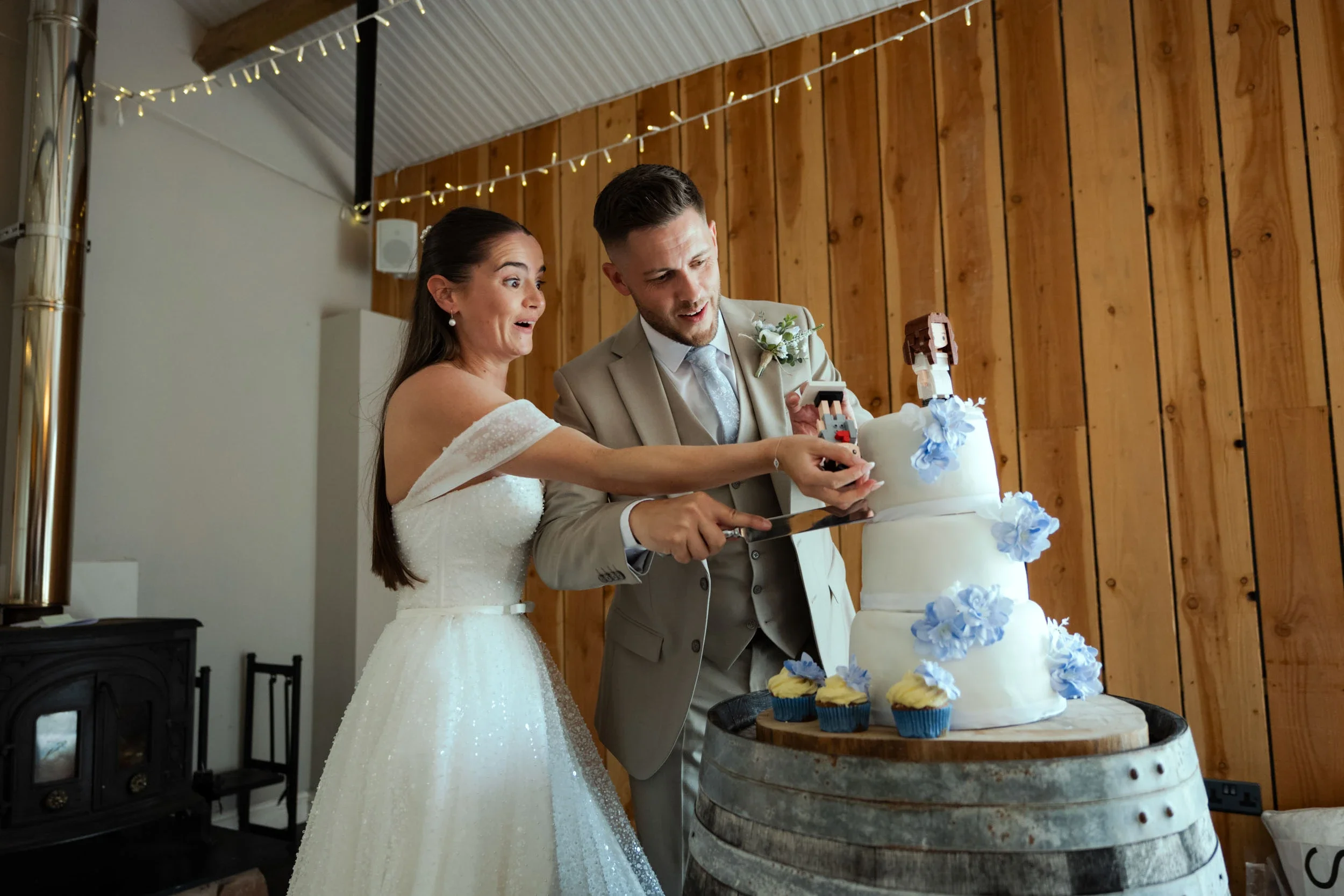 A bride and groom cut a wedding cake together at their wedding reception, with surprised expressions on their faces. The cake is decorated with blue flowers and topped with a figurine. They are standing behind a wooden barrel.
