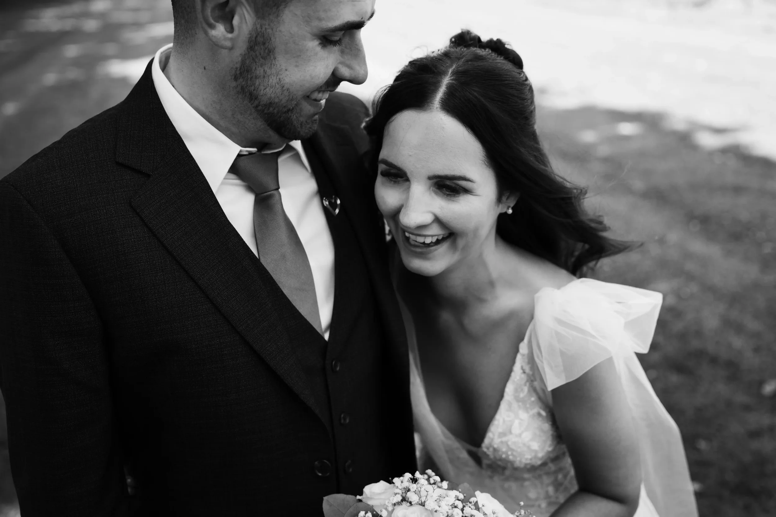 A black and white photo of a smiling bride and groom close together, with the groom in a suit and tie and the bride in a wedding dress holding a bouquet, outdoors near the water.