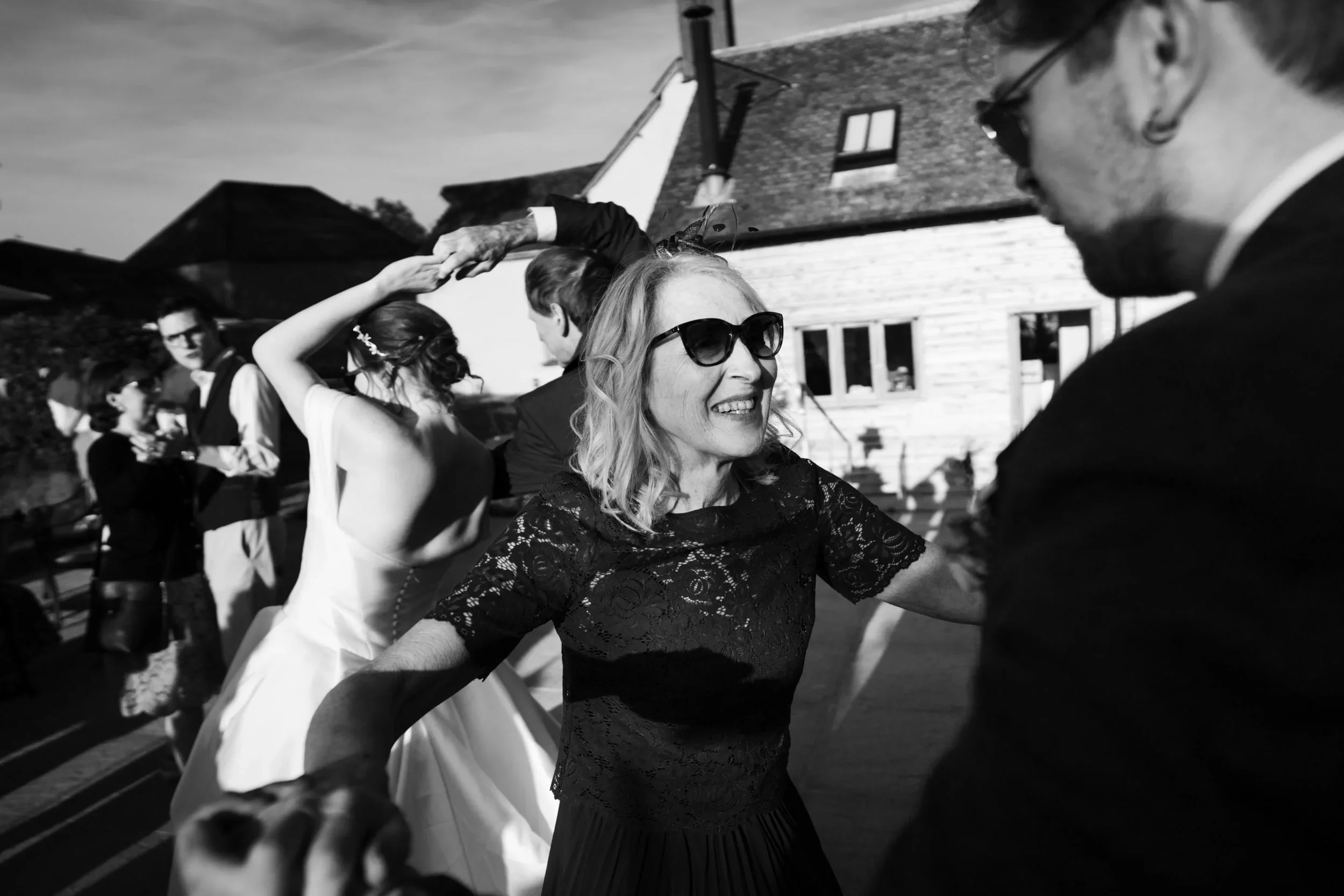 People dancing at an outdoor wedding reception, including a woman in sunglasses and a lace dress smiling and a woman in a white wedding dress.