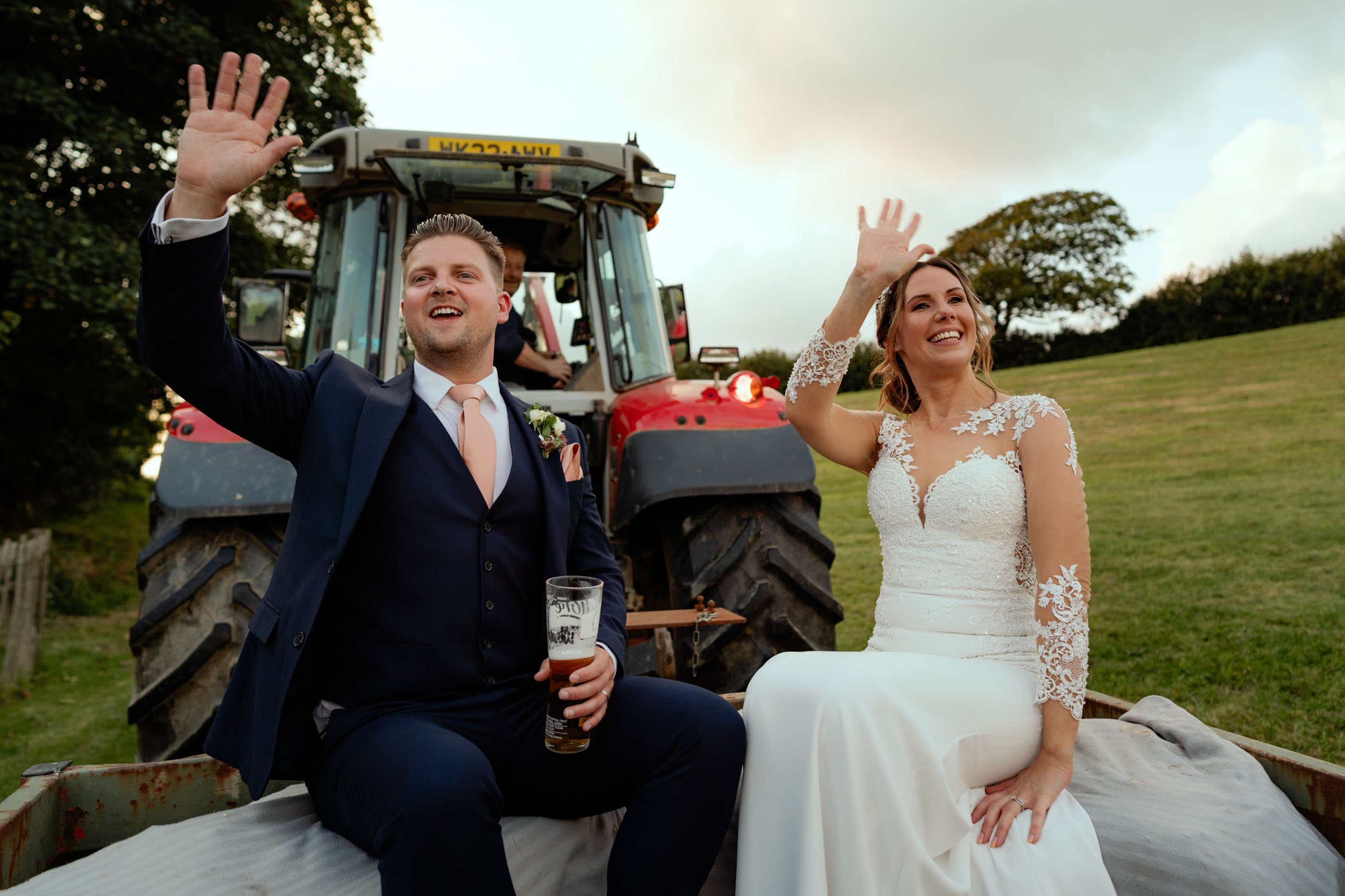 Newlywed couple waving from a tractor on their wedding day, with the groom holding a pint of beer and dressed in a blue suit, while the bride wears a white lace wedding dress, outdoors on a grassy hillside.