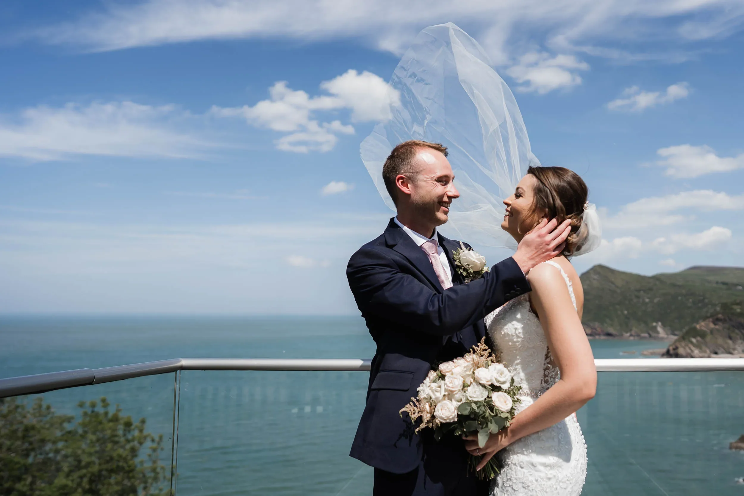 A newlywed couple stands on a balcony overlooking the sea and hills, smiling at each other. The groom is holding the bride's face, and she is holding a bouquet of white roses. The sky is blue with scattered clouds.