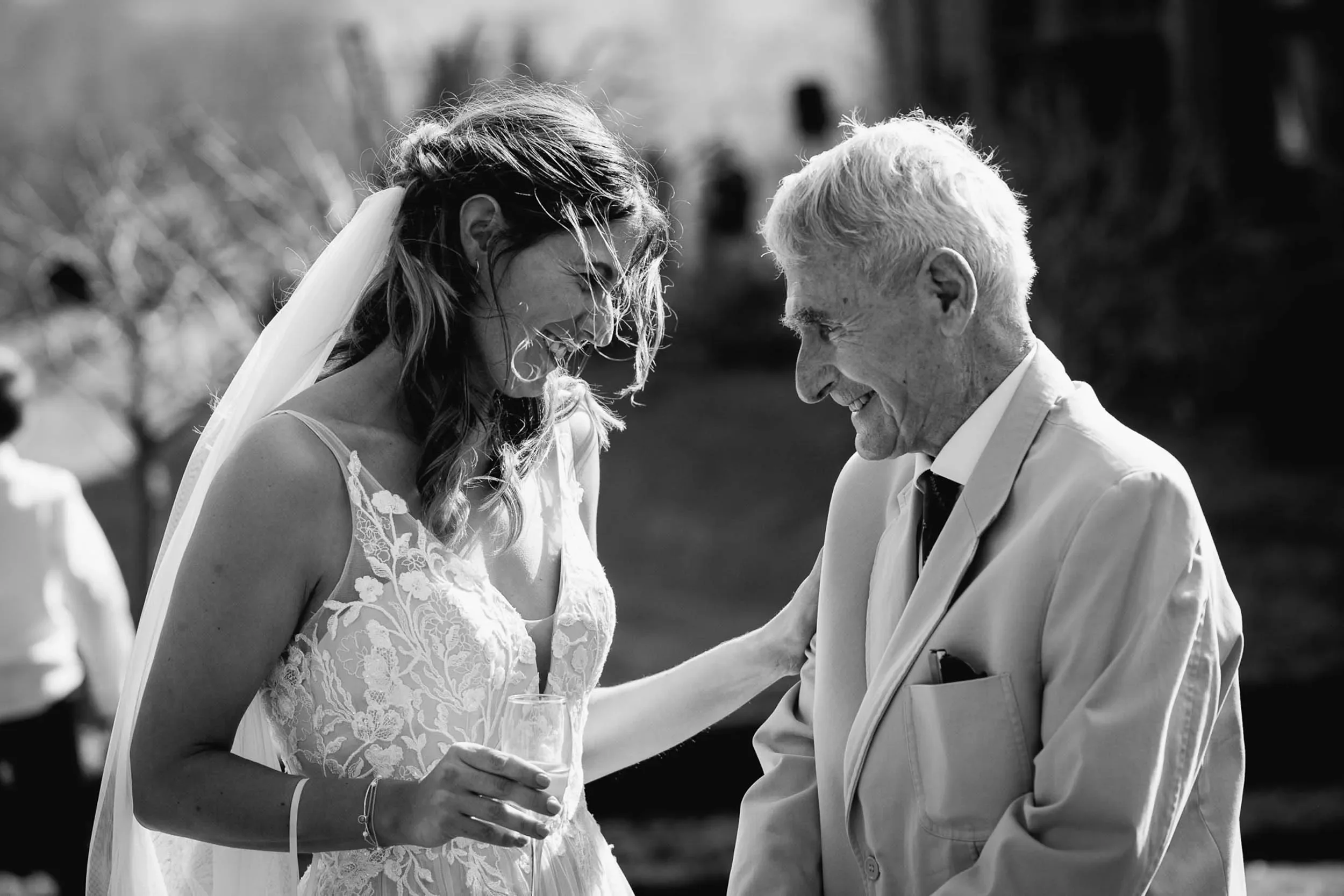 A bride in a lace wedding dress and veil laughing while holding a glass, interacting with an elderly man in a light-colored suit at an outdoor wedding.