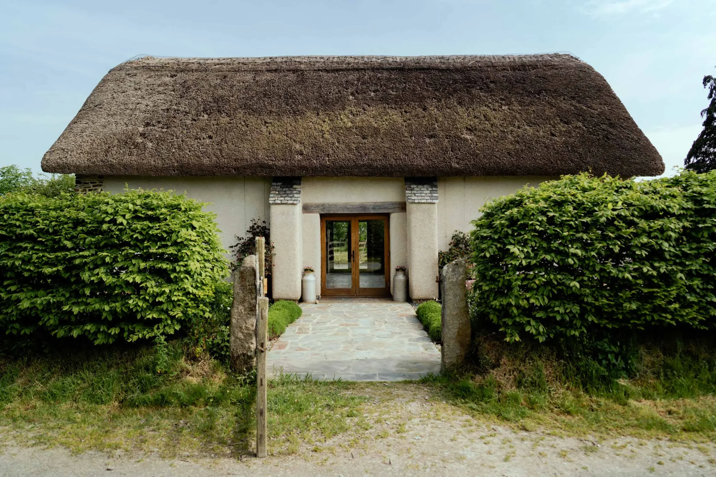 A house with a thatched roof, stone pathway, and green bushes on either side of the entrance, with a wooden door and glass panels.
